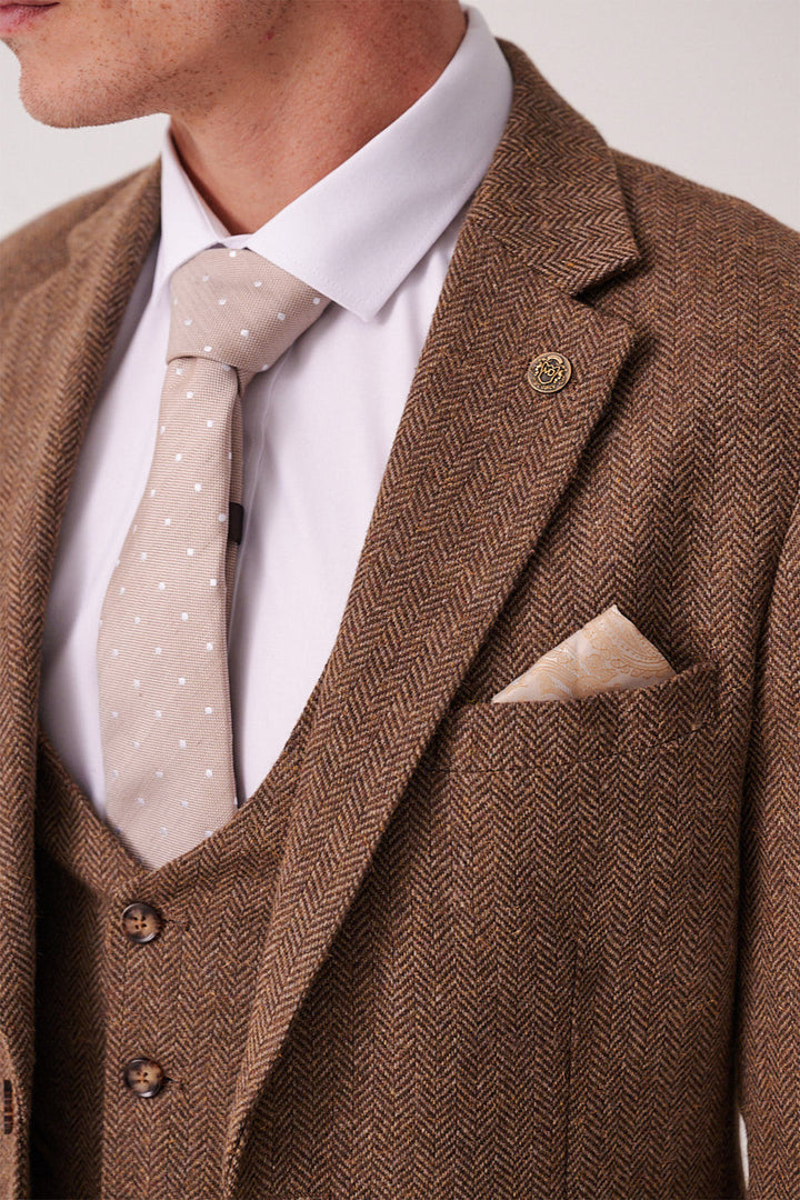 A man wears the marcdarcy DEAN Tan Tweed Three Piece Suit with a beige polka dot tie, white shirt, cream pocket square, and a round lapel pin; only his lower face and upper torso are visible.