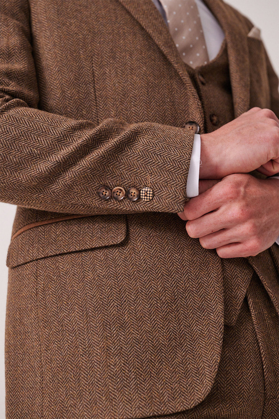 A man adjusts the cuff of the marcdarcy DEAN Tan Tweed Three Piece Suit, featuring brown buttons. He pairs the slim fit jacket with its matching vest, a white shirt, and beige polka dot tie for a refined look.