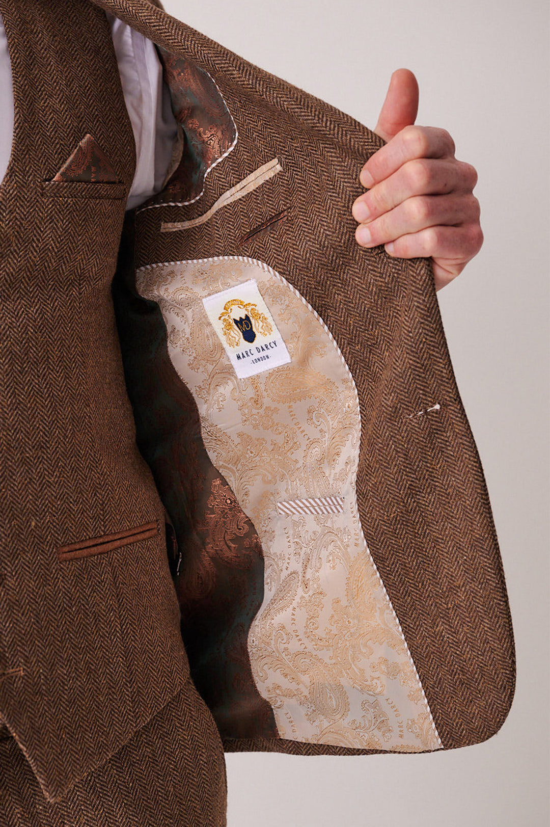 A person holds open a brown herringbone DEAN - Tan Tweed Three Piece Suit by marcdarcy, showing a light paisley lining and a marcdarcy label featuring a lion and crown logo.
