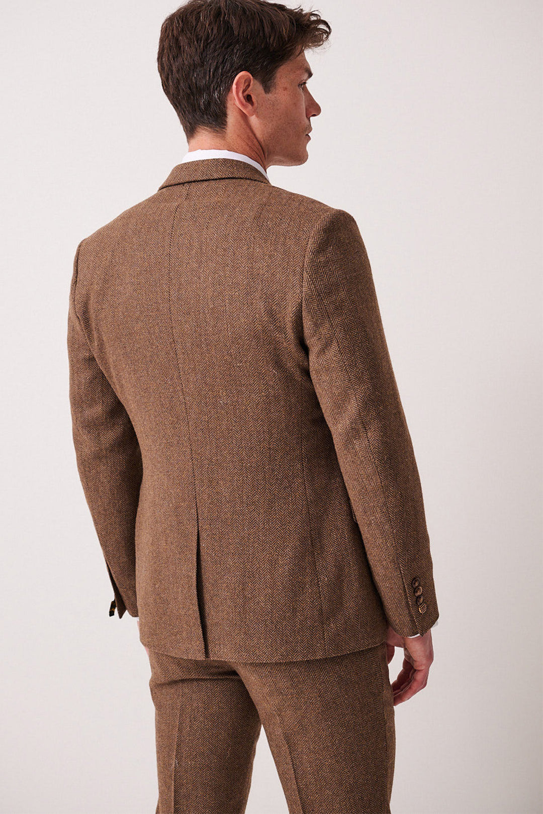 A man stands facing away, wearing the marcdarcy DEAN Tan Tweed Three Piece Suit with matching trousers and a white shirt. He is slightly turned, highlighting the back and side of the suit jacket.