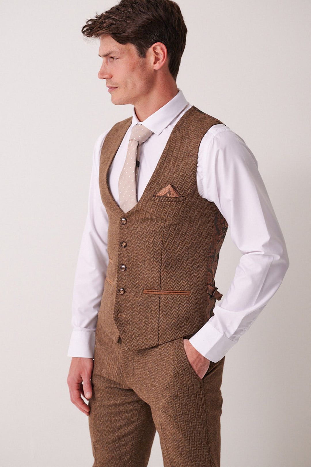 A man faces left against a plain light background, wearing the Marc Darcy DEAN - Tan Tweed Waistcoat, paired with a brown suit, white dress shirt, and beige tie. He has short brown hair and his left hand is in his pocket.
