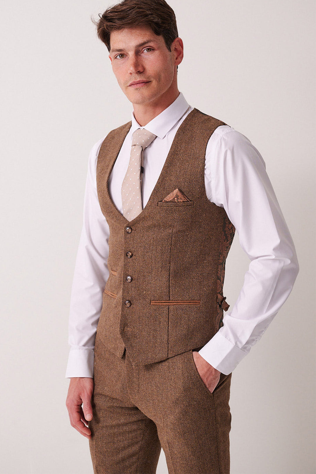 A man stands against a plain light background, wearing the Marc Darcy DEAN - Tan Tweed Waistcoat with matching trousers, a white dress shirt, and a light beige patterned tie.