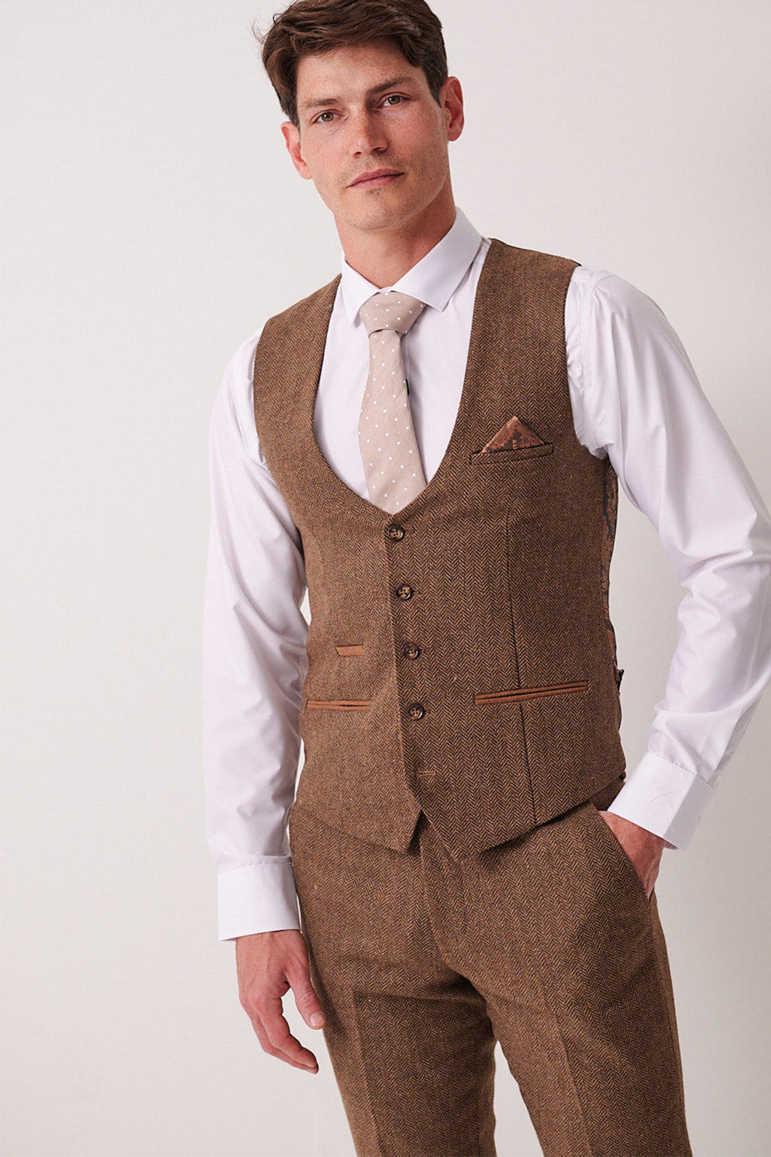 A man wears the Marc Darcy DEAN - Tan Tweed Waistcoat with matching trousers, a white shirt, and beige polka-dot tie, posing against a plain light backdrop with one hand in his pocket, looking at the camera.