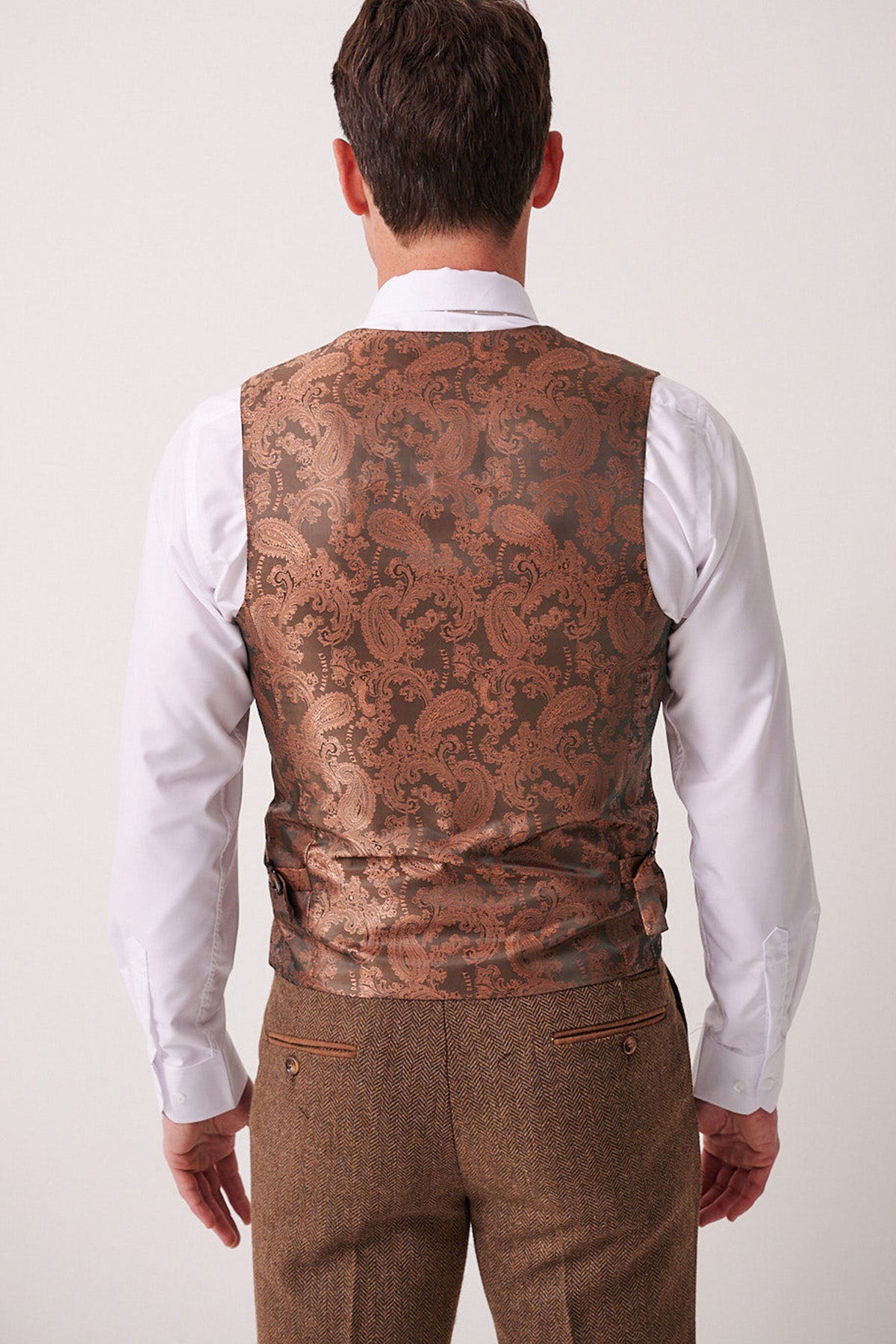 A man stands facing away, wearing a white dress shirt and the Marc Darcy DEAN Tan Tweed Waistcoat, paired with brown textured trousers featuring buttoned back pockets.