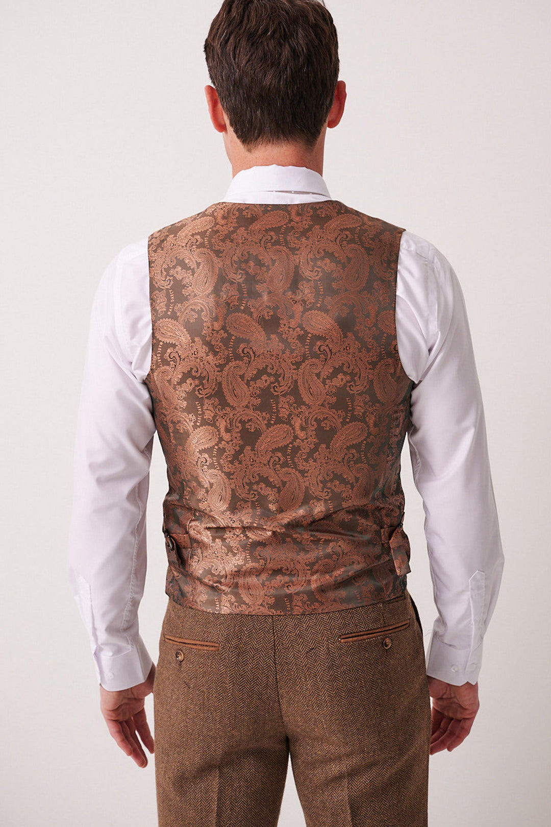 A man seen from behind wears the marcdarcy DEAN Tan Tweed Three Piece Suit, featuring a white shirt and a brown brocade waistcoat with paisley detail. The background is plain and neutral.