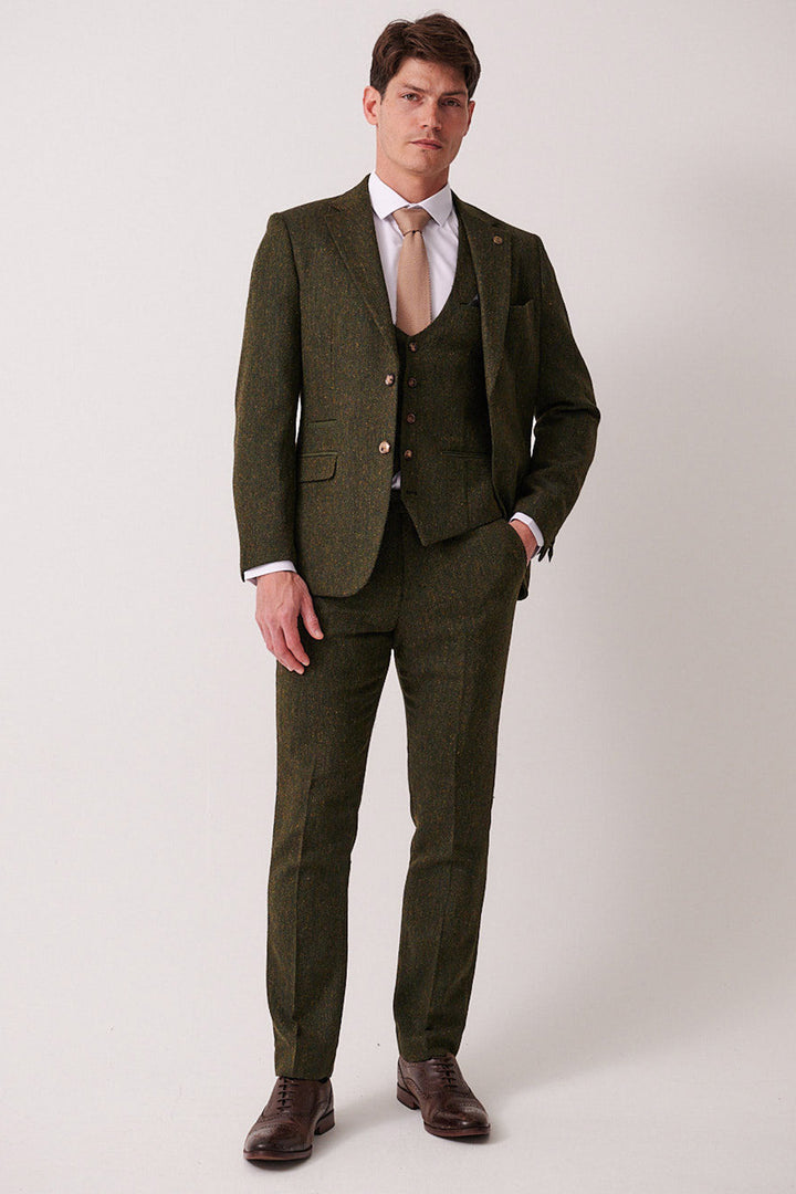 A man with short hair and a neutral expression stands against a plain background, wearing the marcdarcy DEAN Olive Tweed Three Piece Suit with a white shirt, light beige tie, brown dress shoes, and one hand in his pocket.