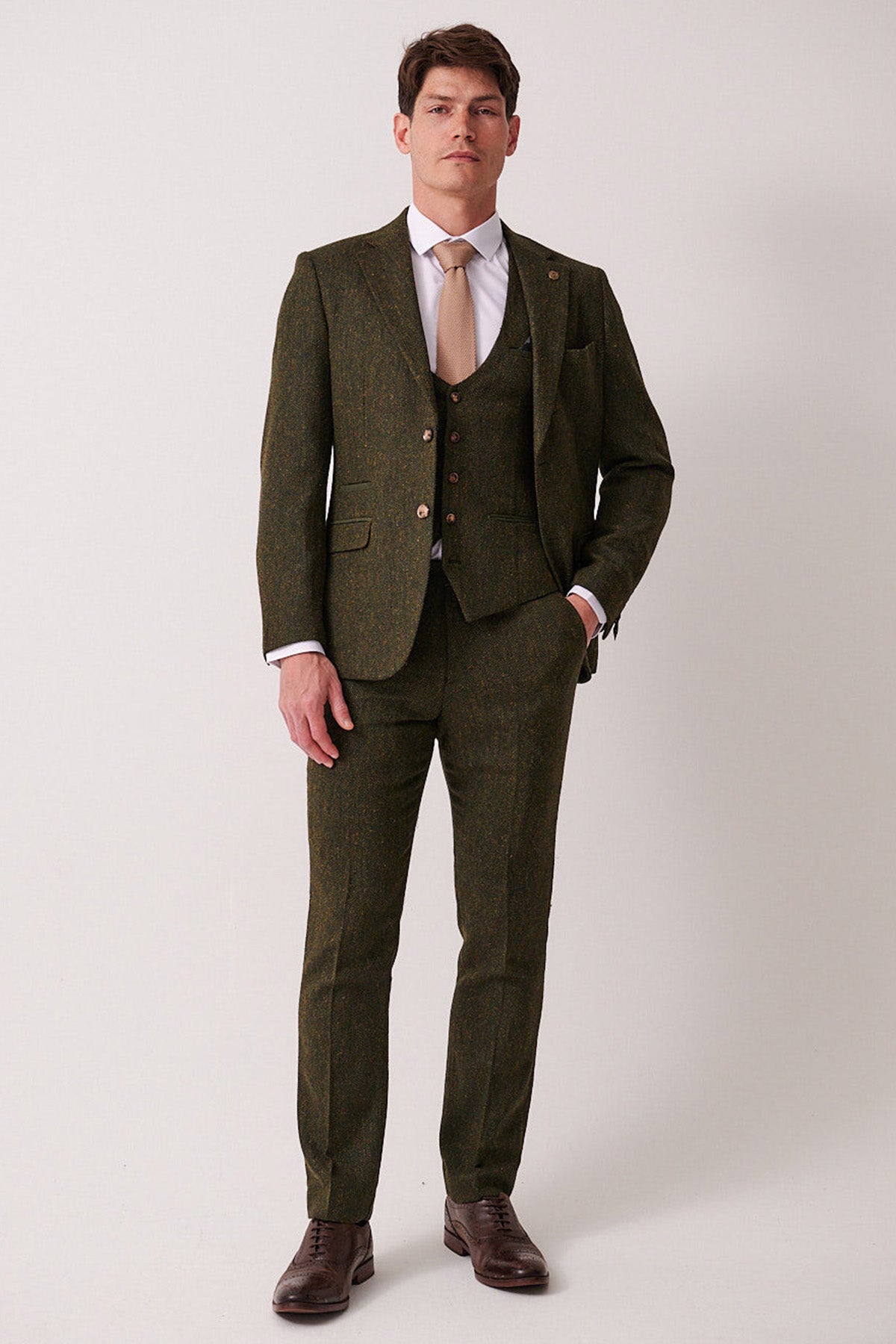 A man faces forward wearing the marcdarcy DEAN Olive Tweed Three Piece Suit—a slim fit wool-blend—paired with a white shirt and tan tie, his left hand in his pocket.