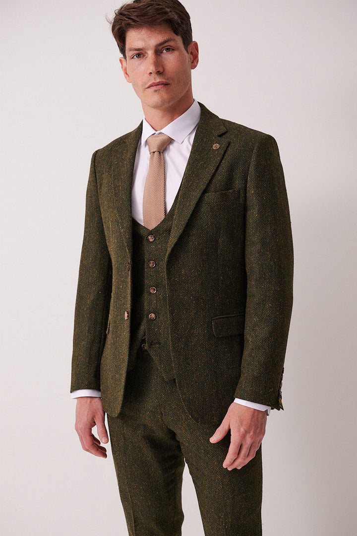 A man wears the marcdarcy DEAN - Olive Tweed Blazer with a white shirt and beige tie, standing against a light background and looking at the camera with a neutral expression.