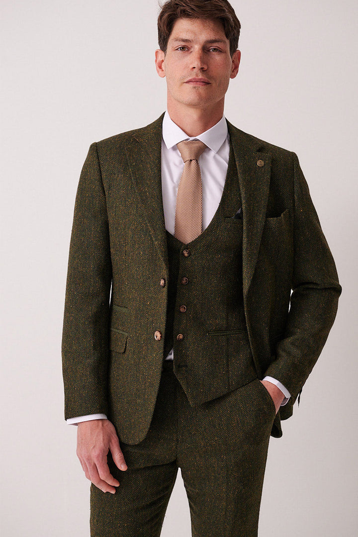 A man faces forward in the marcdarcy DEAN olive tweed blazer, styled as a slim fit three-piece suit with a white dress shirt and beige tie. He smiles slightly, one hand in his pocket, against a simple light background.