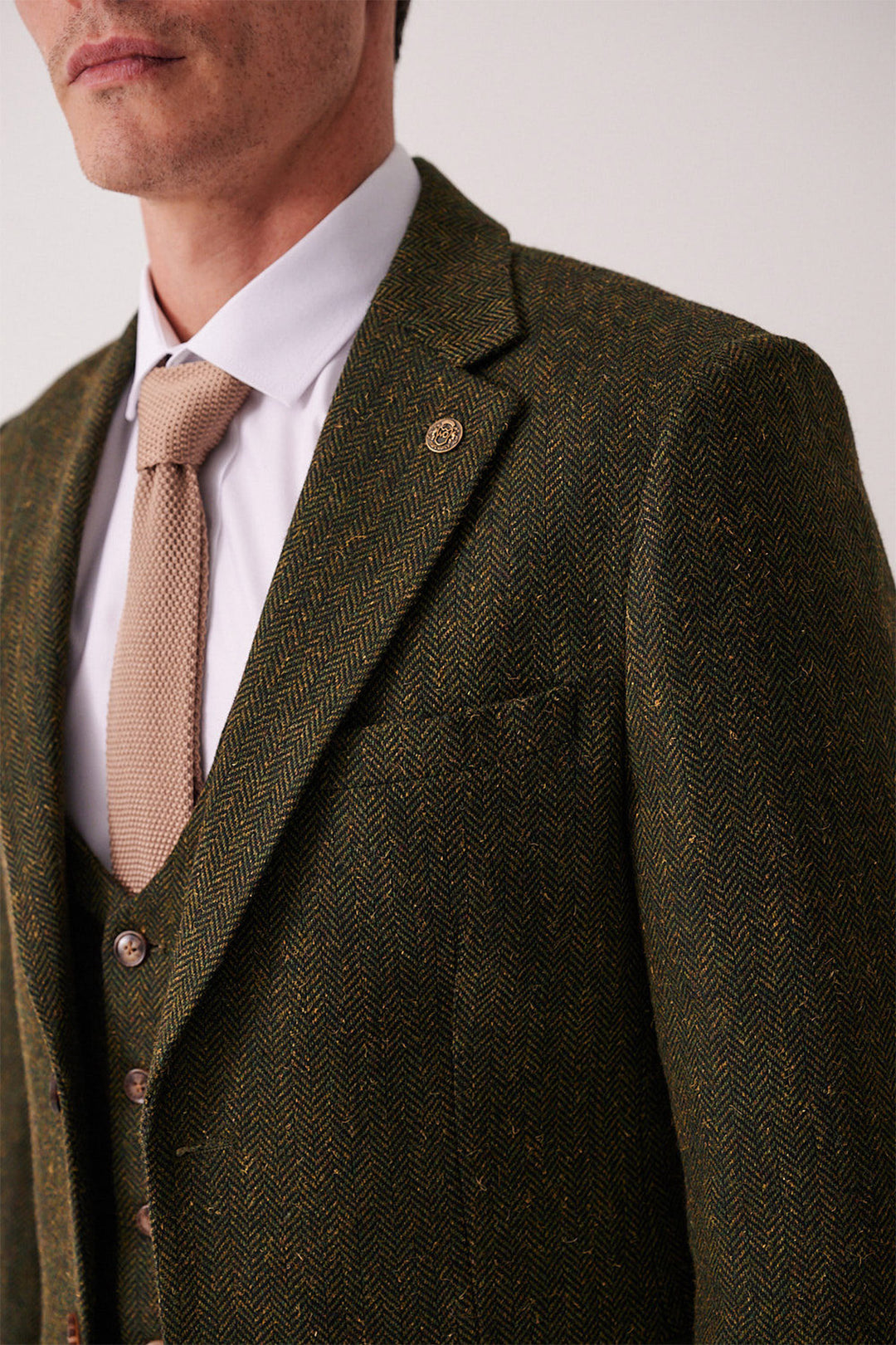 A man wears the marcdarcy DEAN Olive Tweed Blazer, a white dress shirt, and a beige textured tie. The blazer features a lapel pin, and only the lower half of his face is visible.