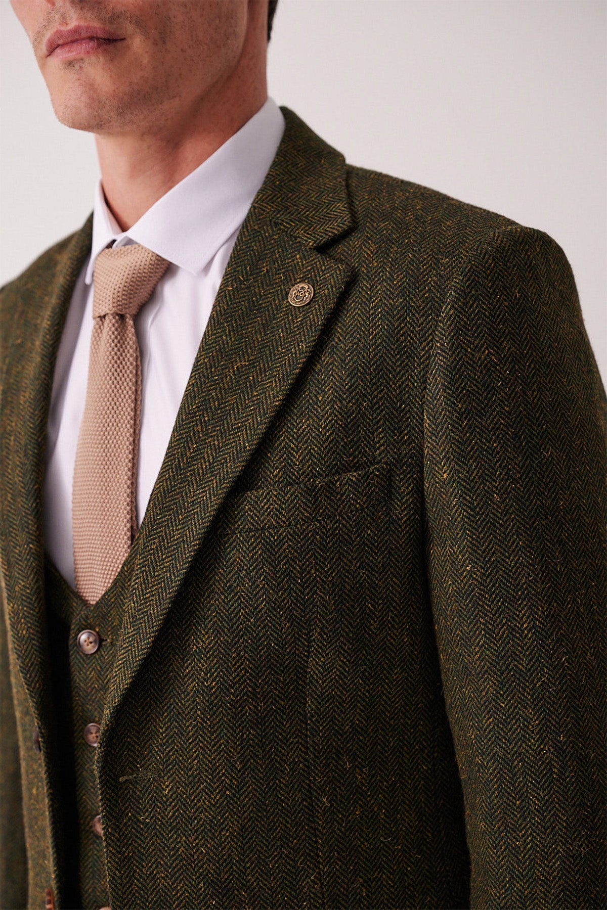 A man wears the marcdarcy DEAN Olive Tweed Blazer, a white dress shirt, and a beige textured tie. The blazer features a lapel pin, and only the lower half of his face is visible.
