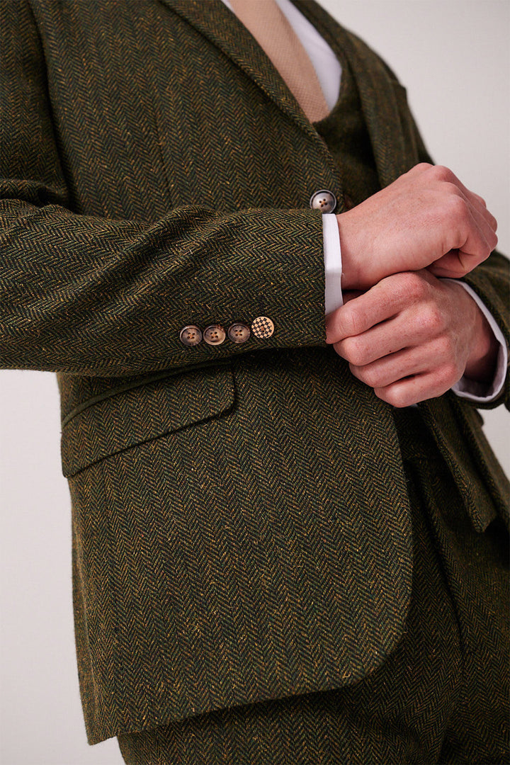 A person in a slim fit marcdarcy DEAN olive tweed blazer adjusts their sleeve to reveal four cuff buttons and a tan tie beneath, highlighting the jacket's textured herringbone detail and stylish features.