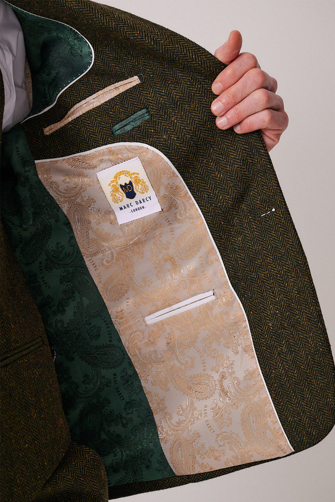 A person opens a DEAN Olive Tweed Three Piece Suit by marcdarcy, showing an ornate beige paisley lining, green pocket, and a white label—an elegant touch in this slim fit men’s tweed ensemble.