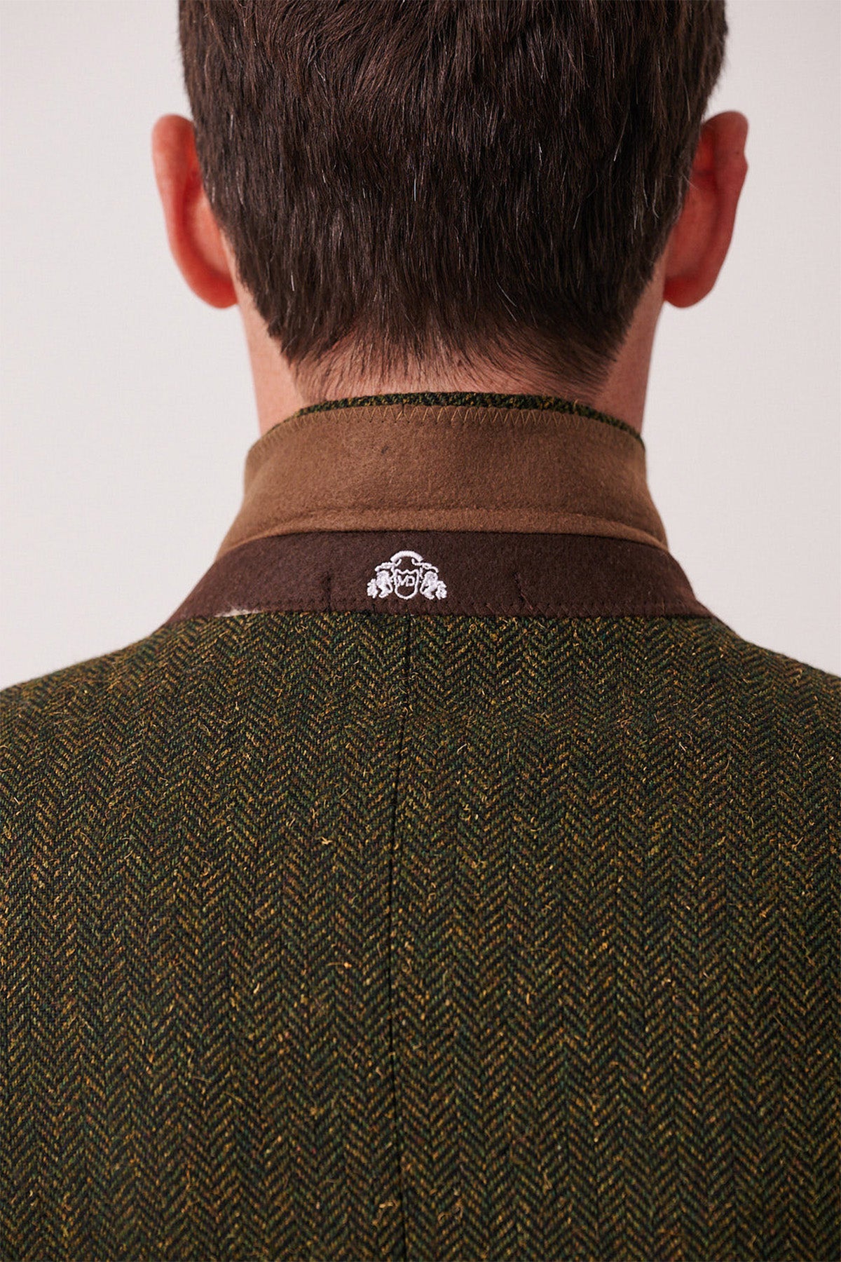 A person shown from behind wears the marcdarcy DEAN - Olive Tweed Blazer, a slim fit men’s blazer with a green and brown herringbone pattern, brown collar, and a small white embroidered logo at the back neck.