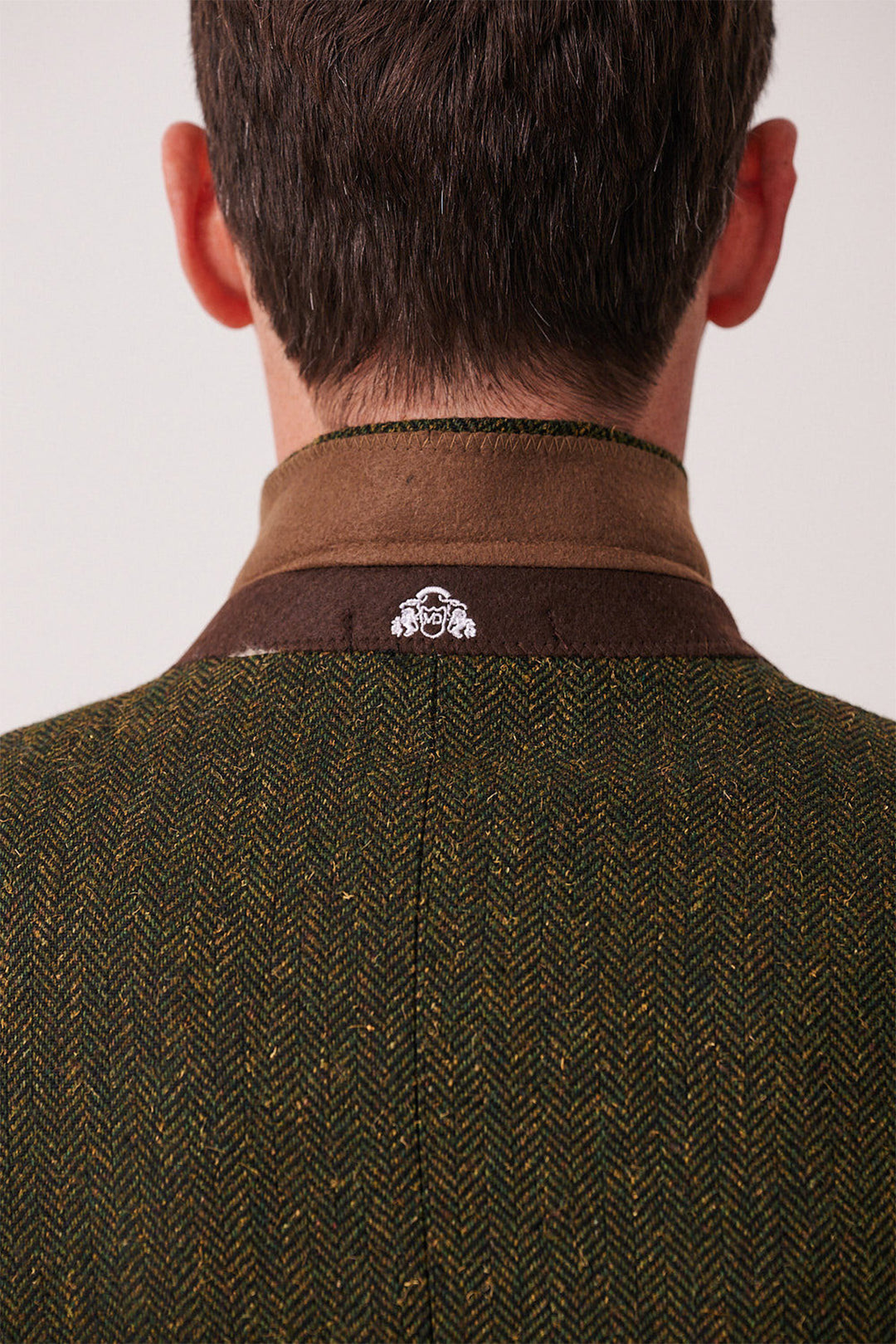 A person seen from behind wears the marcdarcy DEAN - Olive Tweed Three Piece Suit, featuring a slim fit, brown collar, and herringbone pattern with a small white embroidered logo—combining modern style and classic men's tweed details.