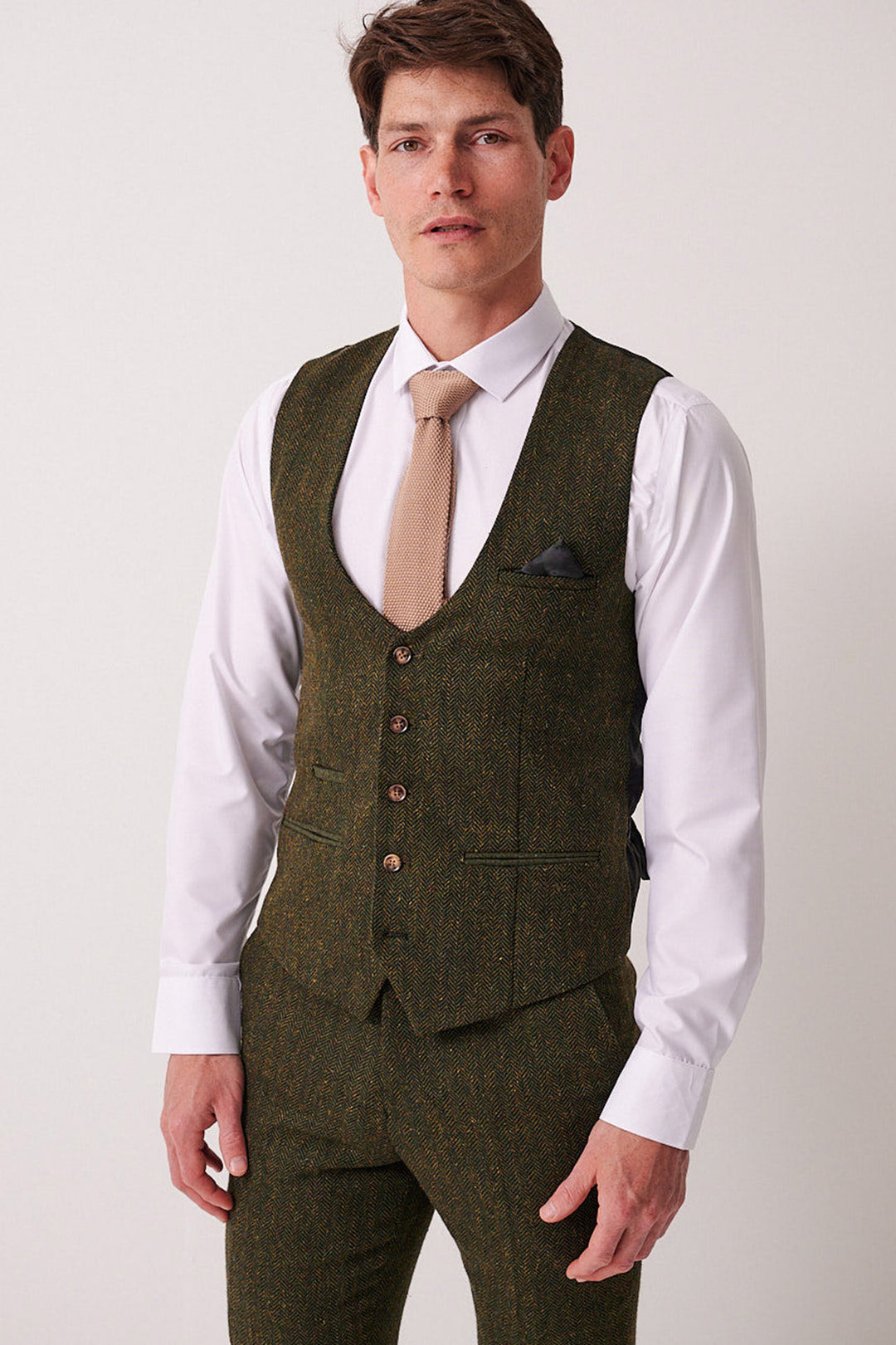 A man wears the Marc Darcy DEAN Olive Tweed Waistcoat with matching trousers, a white dress shirt, and a tan tie, standing facing the camera against a plain light background.