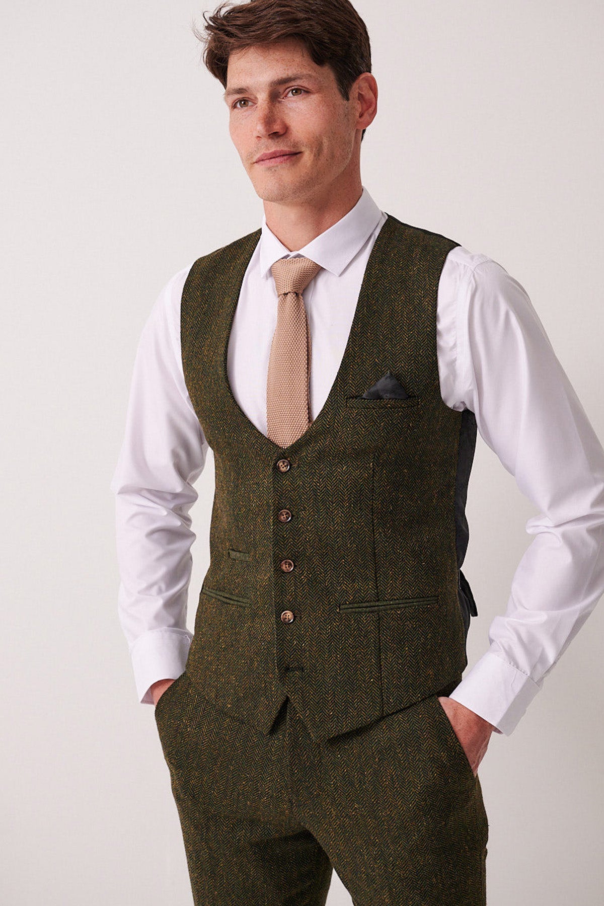 A man stands against a plain background, wearing a white dress shirt, brown tie, and the Marc Darcy DEAN Olive Tweed Waistcoat with matching trousers. His hands in his pockets, he gazes to the side with a slight smile.