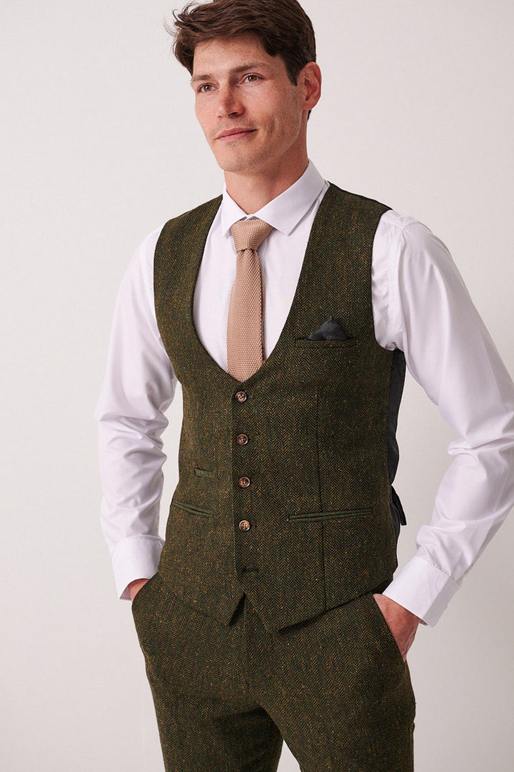 A man stands against a plain background, wearing a white dress shirt, brown tie, and the Marc Darcy DEAN Olive Tweed Waistcoat with matching trousers. His hands in his pockets, he gazes to the side with a slight smile.