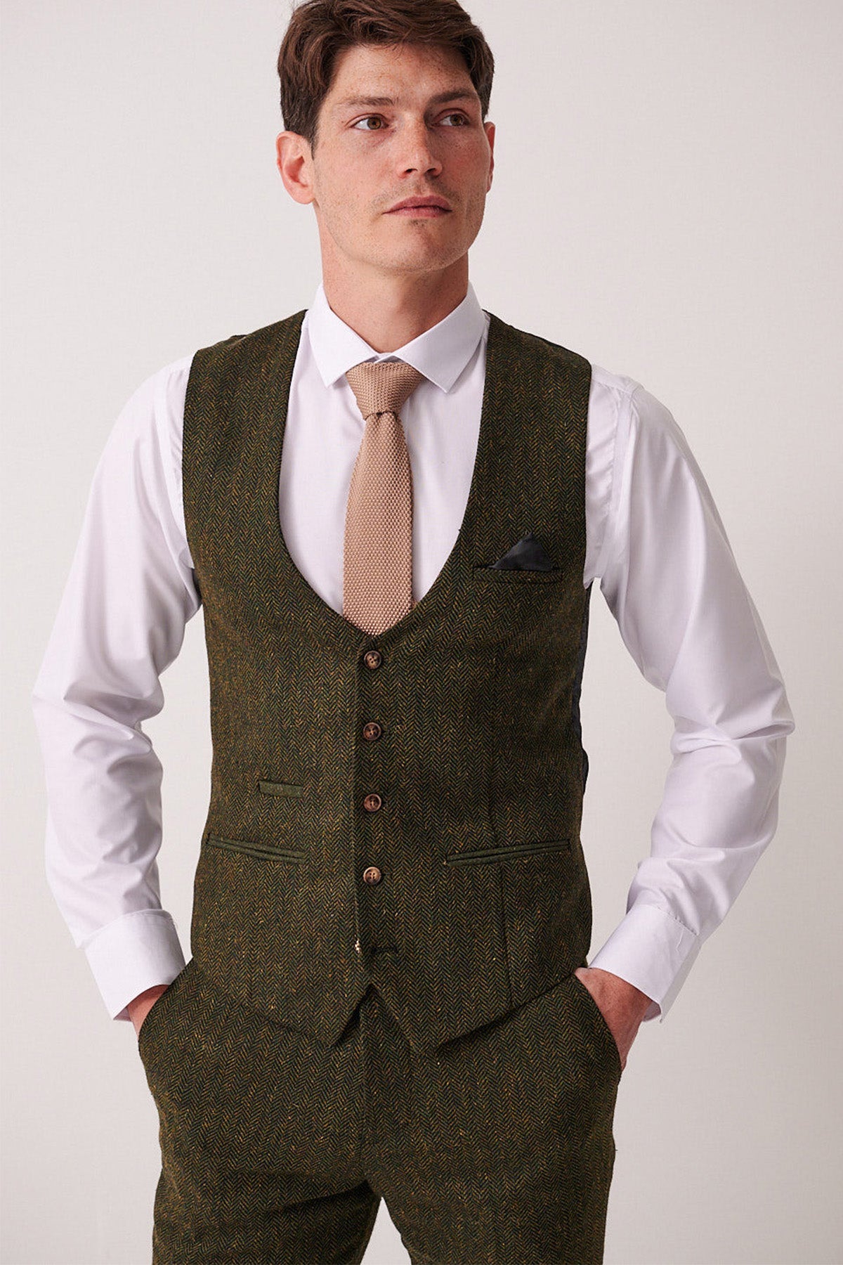 A man wearing a white shirt, tan tie, and the Marc Darcy DEAN - Olive Tweed Waistcoat with brown buttons stands with hands in his pockets, looking to the side against a plain background.
