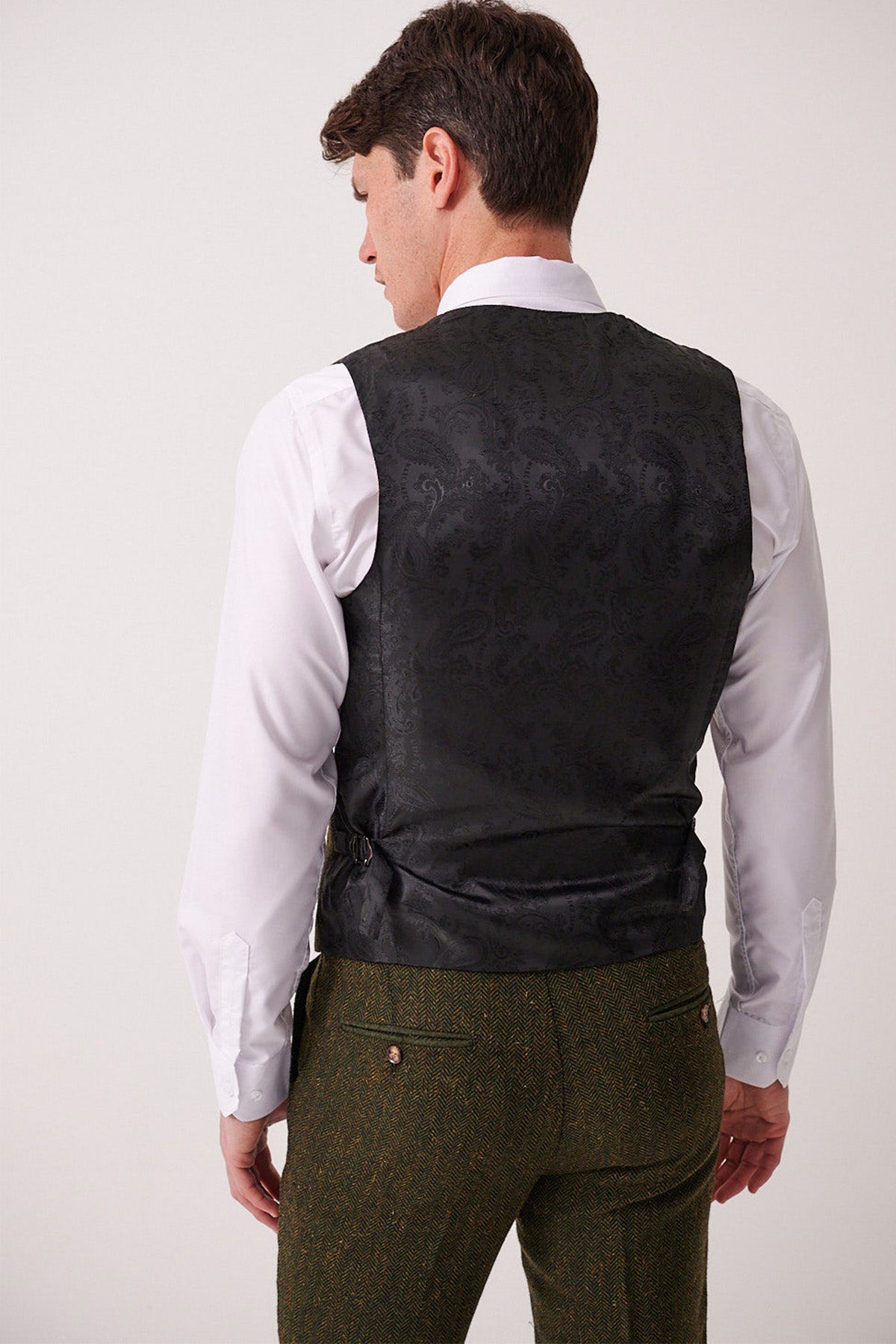 A man stands facing away against a plain light background, wearing a white dress shirt and the Marc Darcy DEAN Olive Tweed Waistcoat, paired with tweed trousers featuring buttoned back pockets.