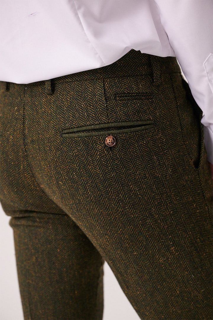 Close-up of a person wearing marcdarcy's DEAN olive tweed trousers. The image highlights the slim fit men's herringbone back pocket with a button closure, paired with a white dress shirt.