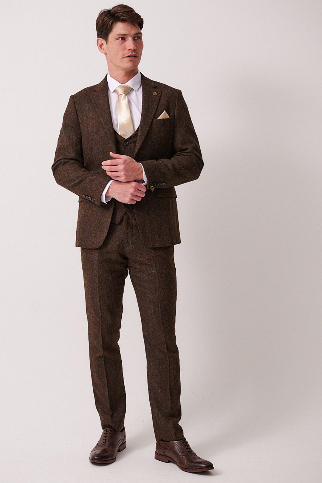 A man with short brown hair stands against a plain light background, wearing the marcdarcy DEAN Brown Tweed Three Piece Suit with a white shirt, beige tie and pocket square, and brown dress shoes, looking slightly to the side.