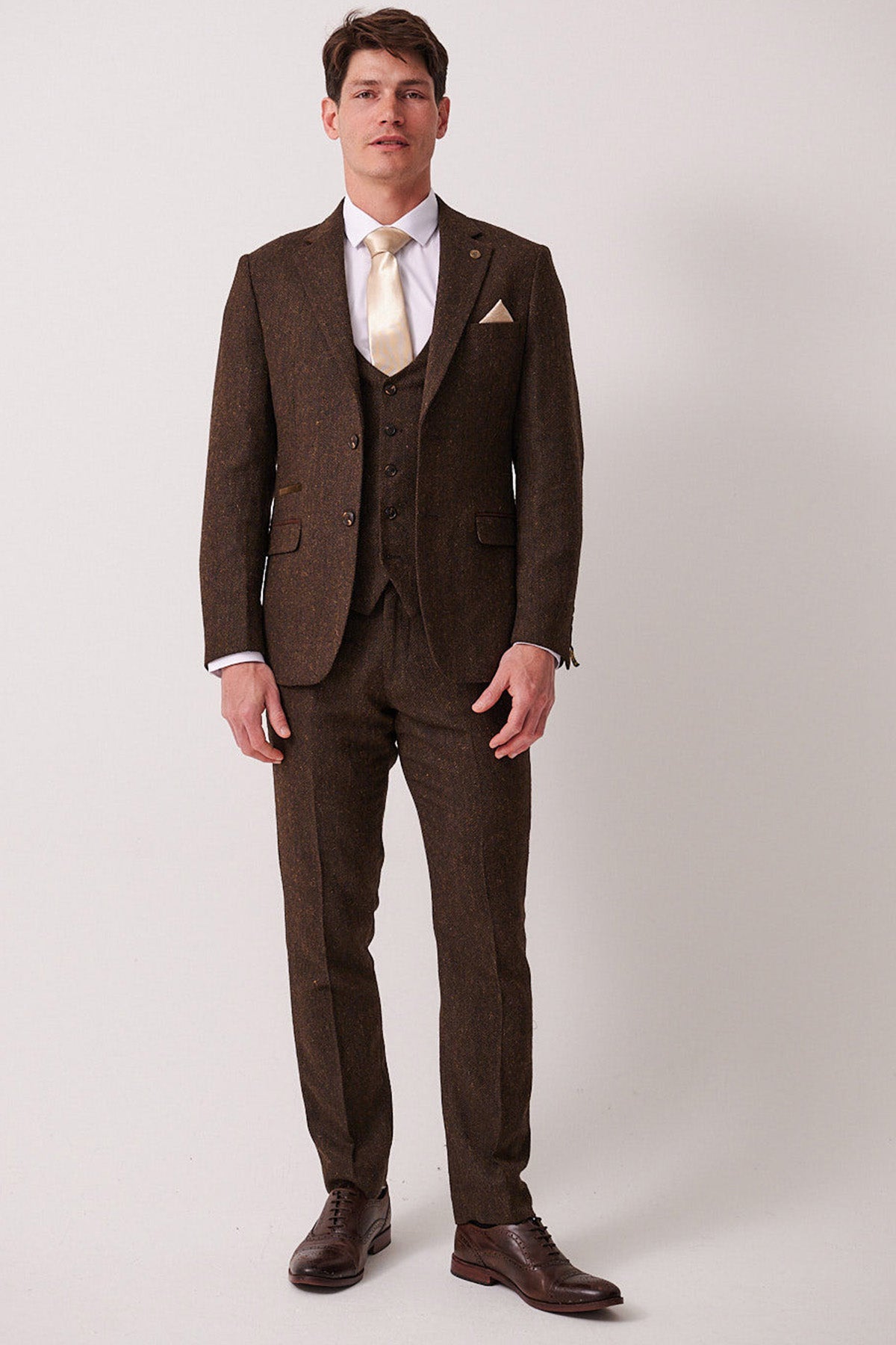 A man with short brown hair and a neutral expression stands against a plain light background, wearing the marcdarcy DEAN Brown Tweed Three Piece Suit with a white dress shirt, cream tie, and brown dress shoes.