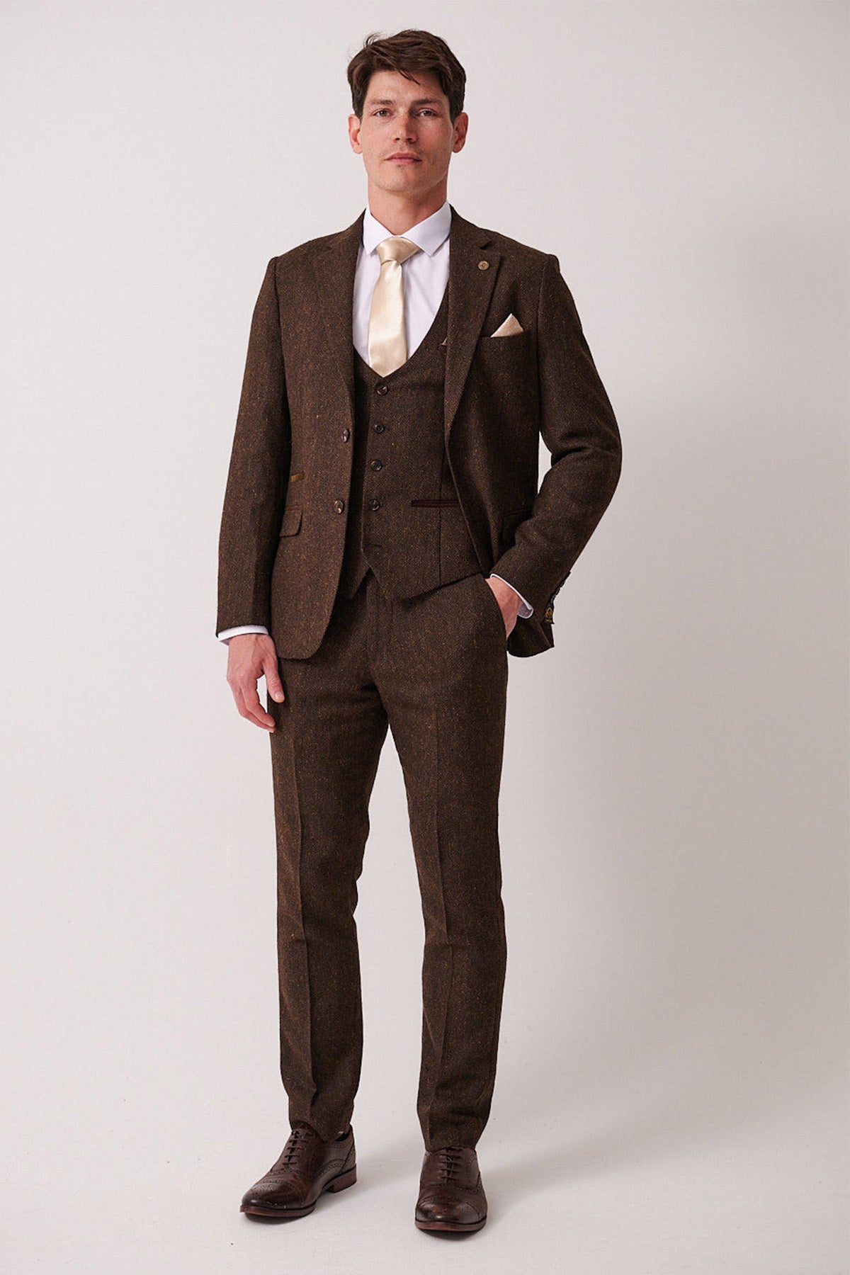 A man stands against a plain light background, wearing the marcdarcy DEAN Brown Tweed Three Piece Suit with a matching vest, white shirt, cream tie, pocket square, and brown dress shoes. He has one hand in his pocket and faces forward.