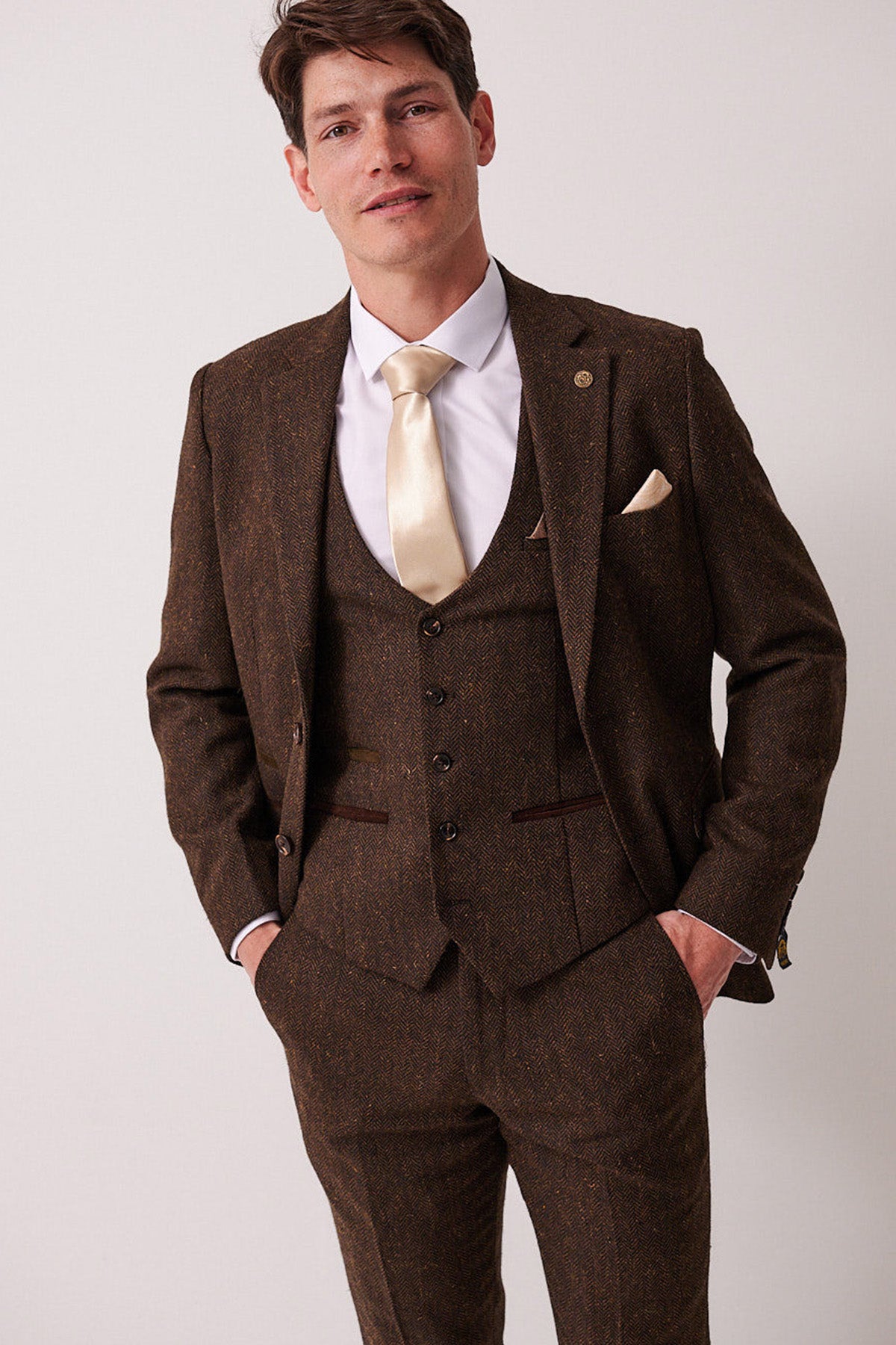 A man stands facing the camera against a plain light background, wearing the marcdarcy DEAN Brown Tweed Blazer, styled with a white dress shirt and beige tie, his hands in his pockets.