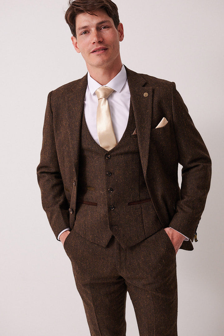 A man stands facing the camera against a plain light background, wearing the marcdarcy DEAN Brown Tweed Blazer, styled with a white dress shirt and beige tie, his hands in his pockets.