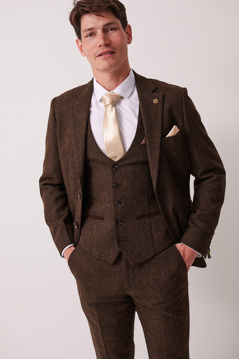 A man wears the marcdarcy DEAN Brown Tweed Three Piece Suit with a herringbone blazer, white shirt, cream satin tie, and pocket square, hands in pockets against a light grey background.