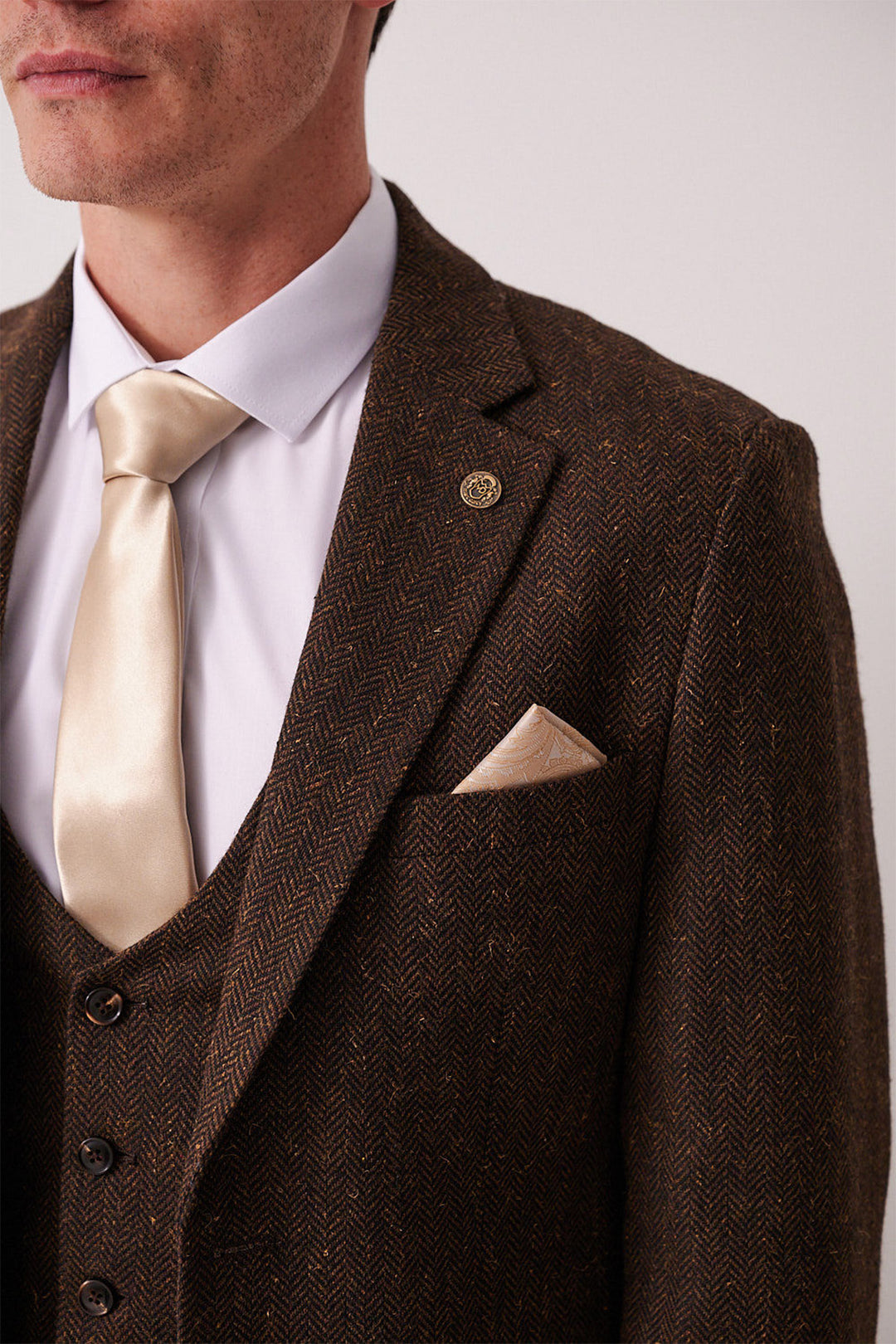 A man wears the marcdarcy DEAN Brown Tweed Three Piece Suit with a white shirt, cream satin tie, and matching pocket square. A cropped photo highlights a lapel pin on the slim fit blazer, capturing its classic elegance.