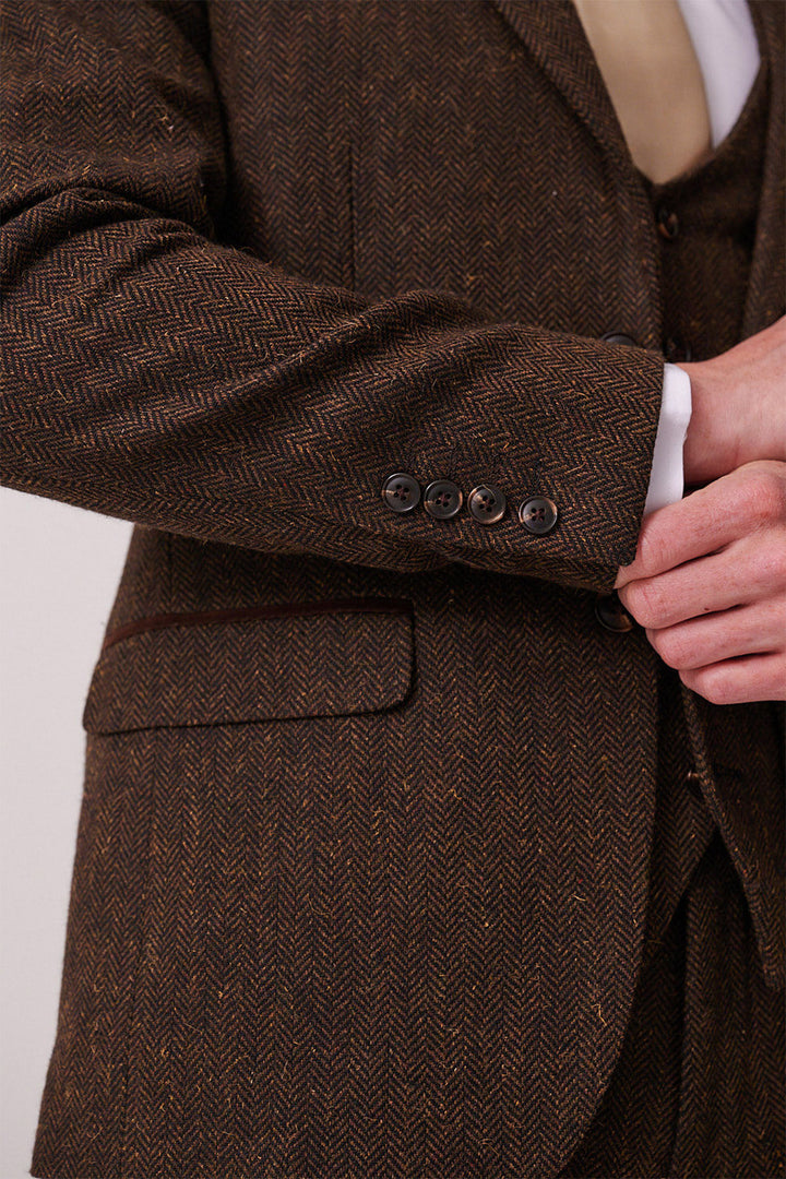 Close-up of a person wearing the marcdarcy DEAN Brown Tweed Three Piece Suit, featuring a herringbone pattern, four dark cuff buttons, beige tie, white shirt, and adjusting the jacket.