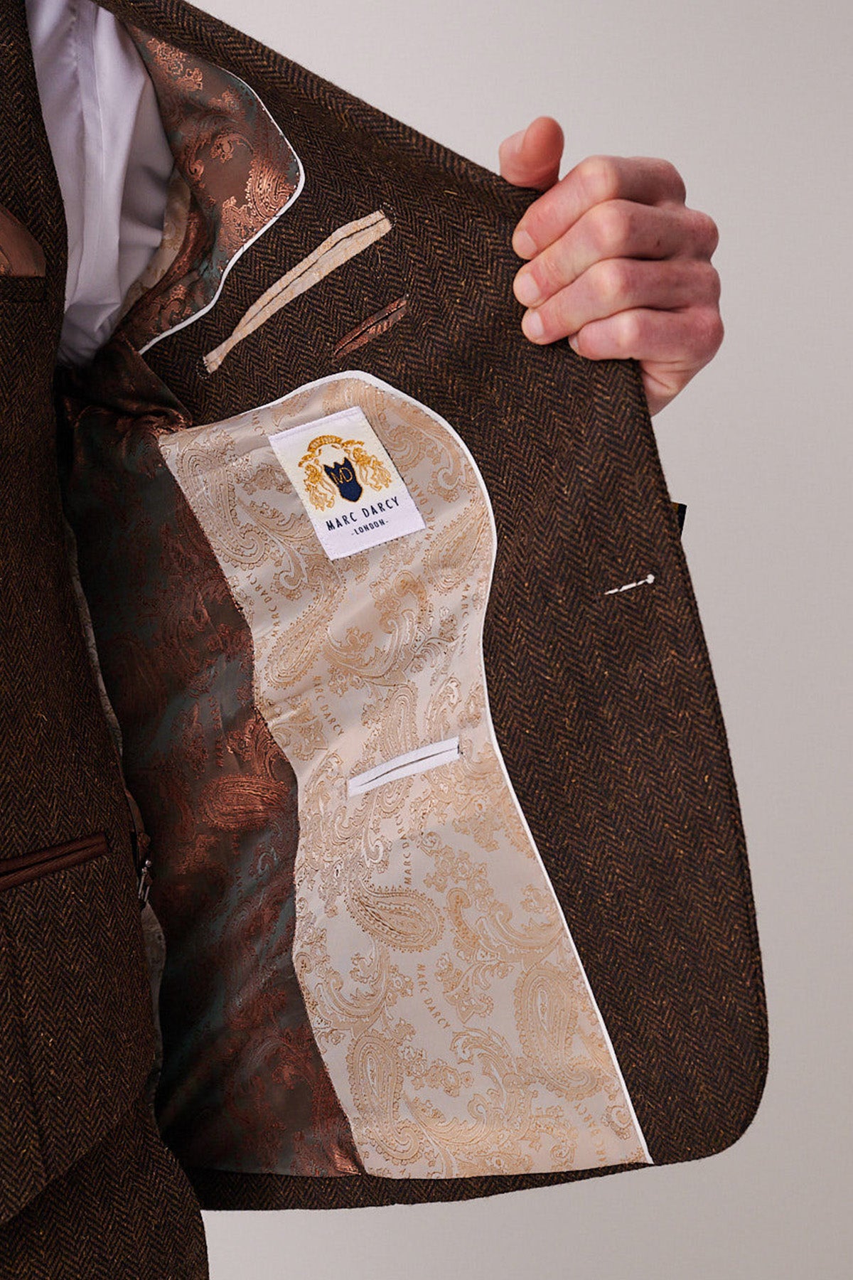 A person opens a brown herringbone suit jacket to reveal an ornate gold paisley lining and a marcdarcy label—a refined touch that pairs perfectly with the DEAN Brown Tweed Blazer.