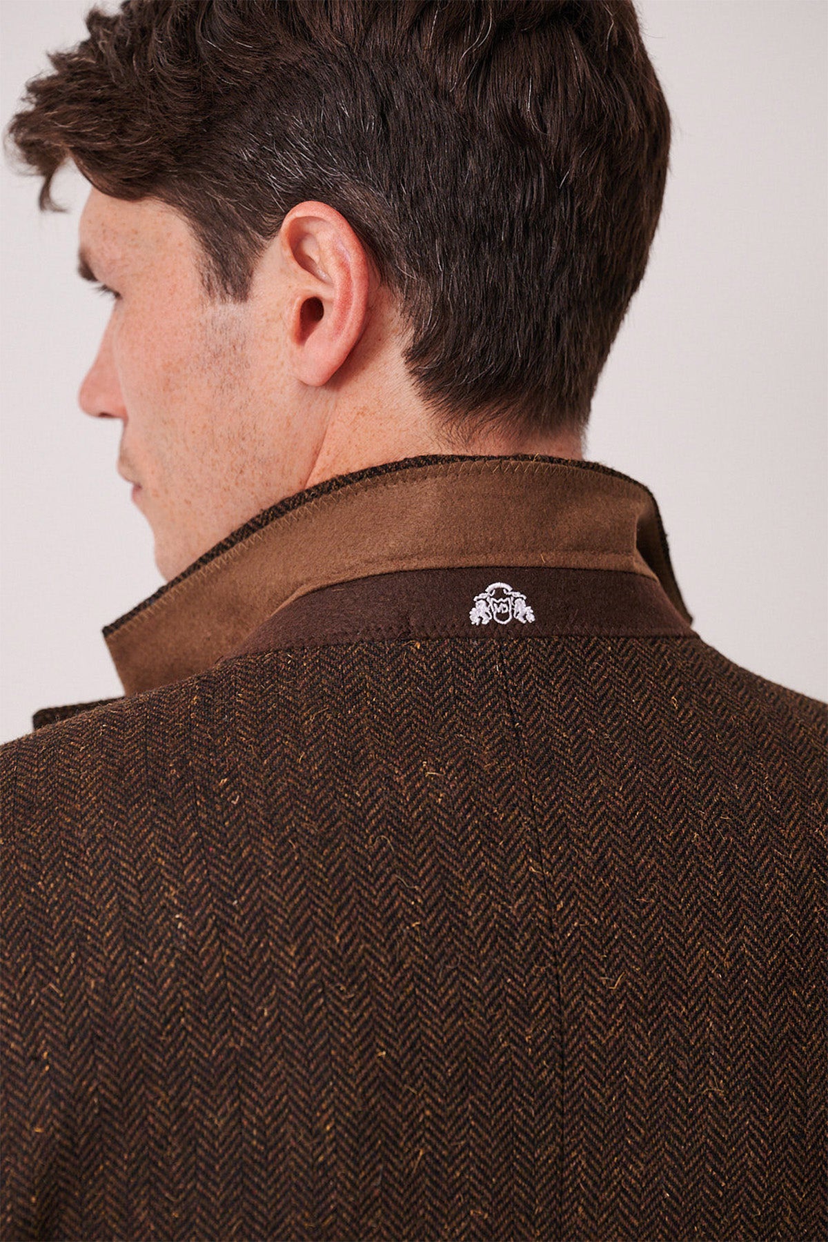 A man with short brown hair is shown from behind, wearing the marcdarcy DEAN Brown Tweed Blazer with its collar turned up. A small white embroidered logo appears on the blazer's collar.