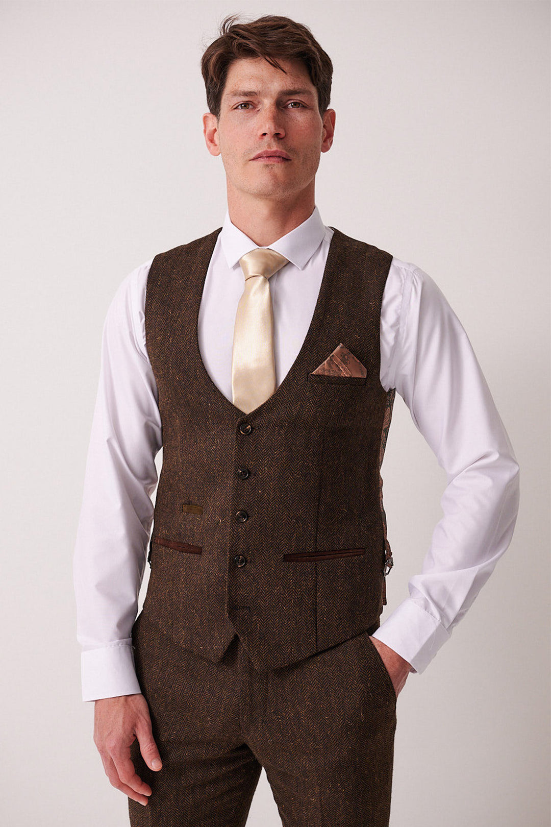 A man wearing the marcdarcy DEAN - Brown Tweed Three Piece Suit, featuring a matching vest and pants, a white dress shirt, and a gold tie stands against a plain light background with one hand in his pocket.