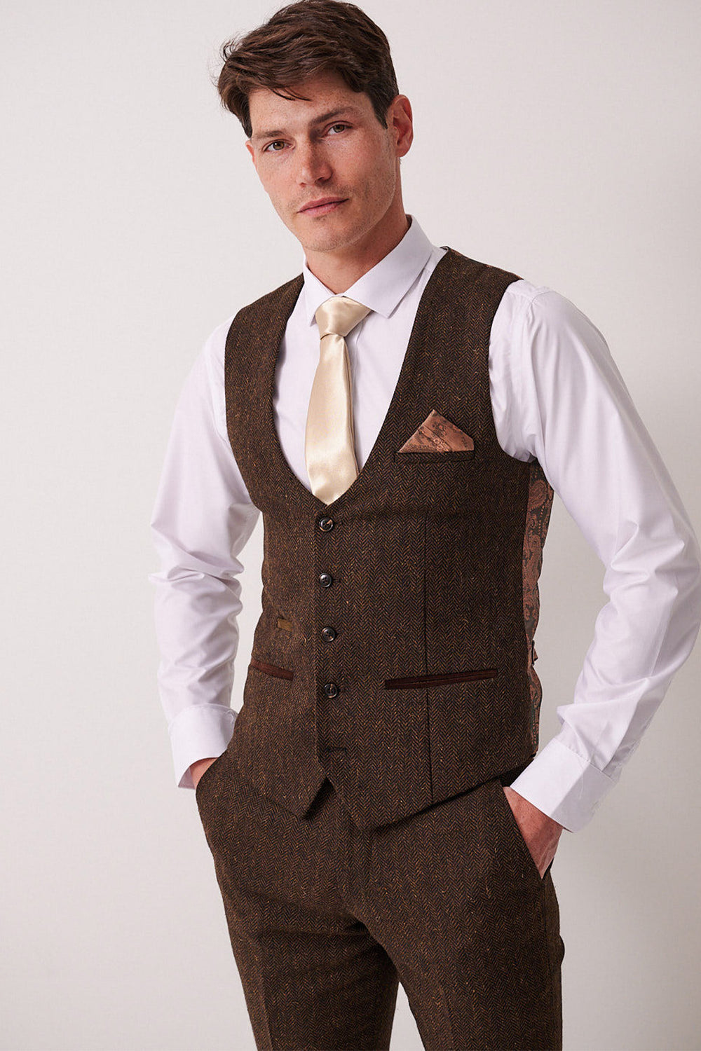 A man with short brown hair stands against a plain background wearing the Marc Darcy DEAN - Brown Tweed Waistcoat, matching trousers, a white shirt, beige tie, and brown pocket square, with his hands in his pockets.
