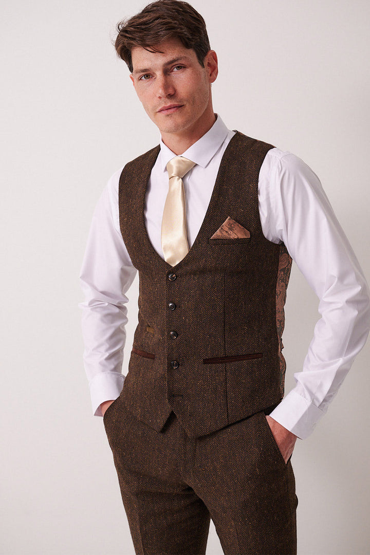 A man wears the Marc Darcy DEAN brown tweed waistcoat over a white shirt and golden tie, posing with his hands in his pockets.