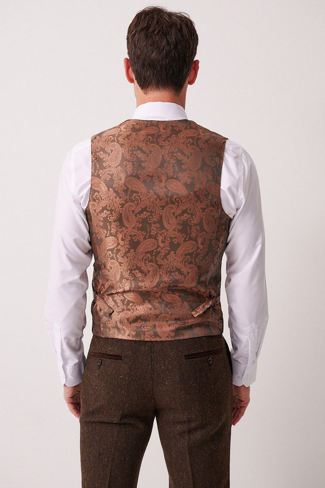 A man faces away from the camera wearing the marcdarcy DEAN Brown Tweed Three Piece Suit. The suit features a white dress shirt, brown waistcoat, and matching trousers against a plain, neutral background.