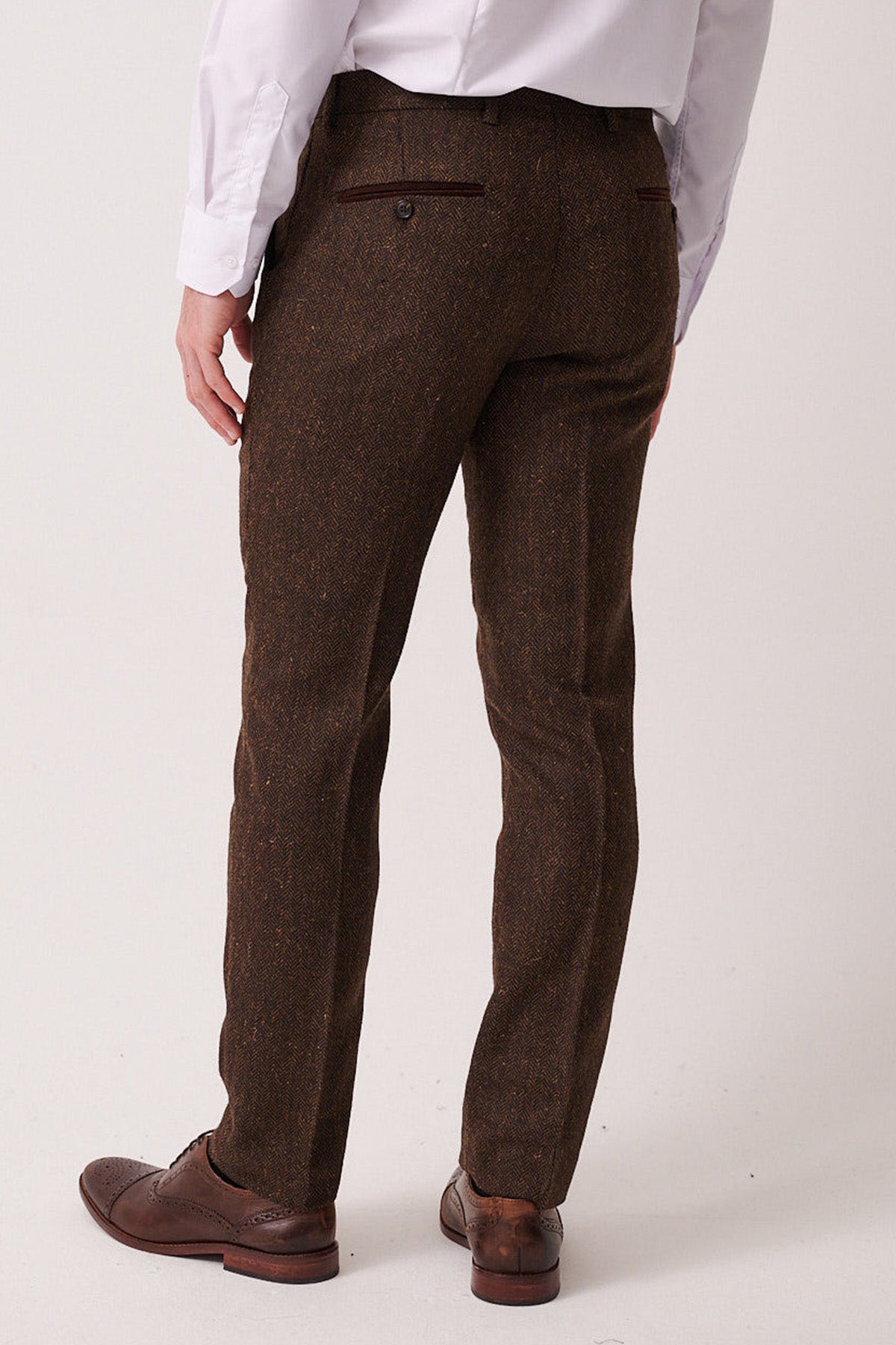 A man in a marcdarcy DEAN Brown Tweed Three Piece Suit, featuring Prince of Wales check, with a white shirt and brown dress shoes stands with his back to the camera against a light background.