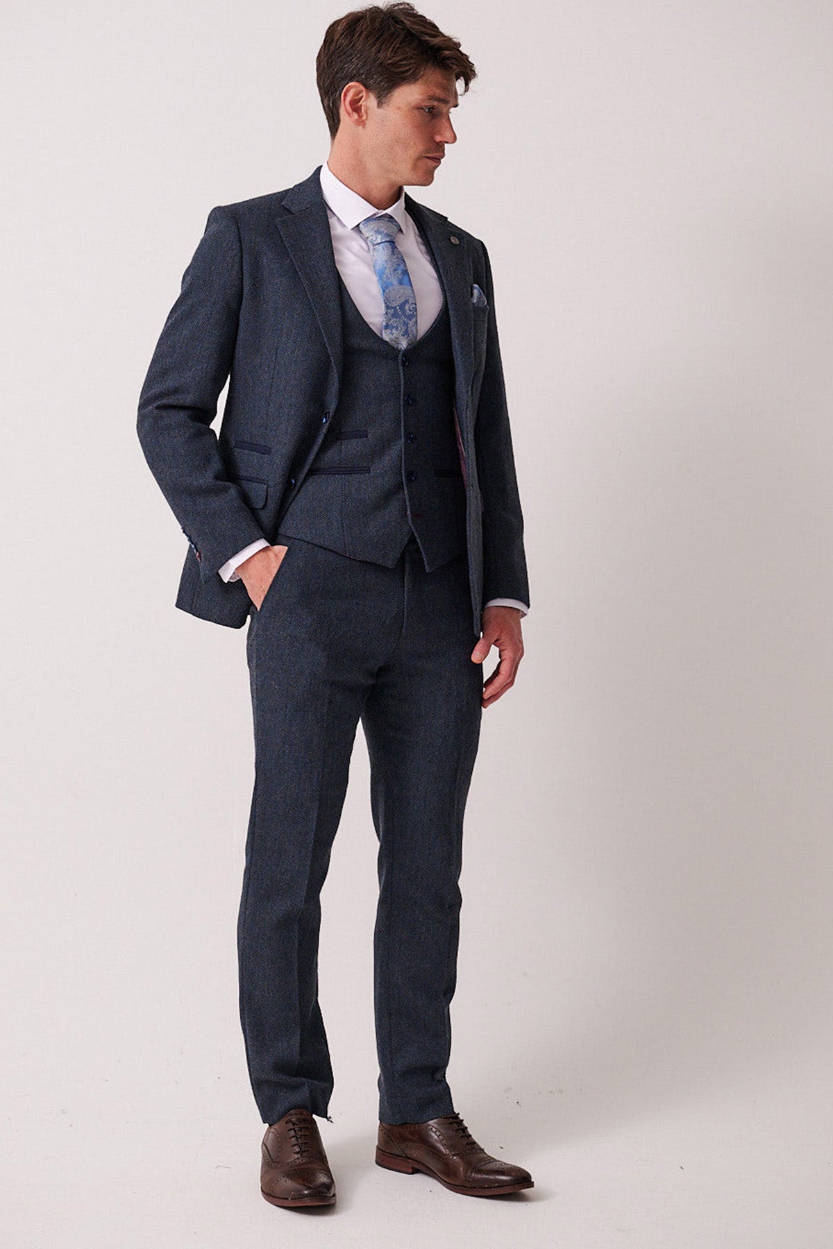 A man in the marcdarcy DEAN Blue Tweed Three Piece Suit with a white shirt, blue patterned tie, and brown dress shoes stands against a plain light background, looking left with one hand in his pocket.