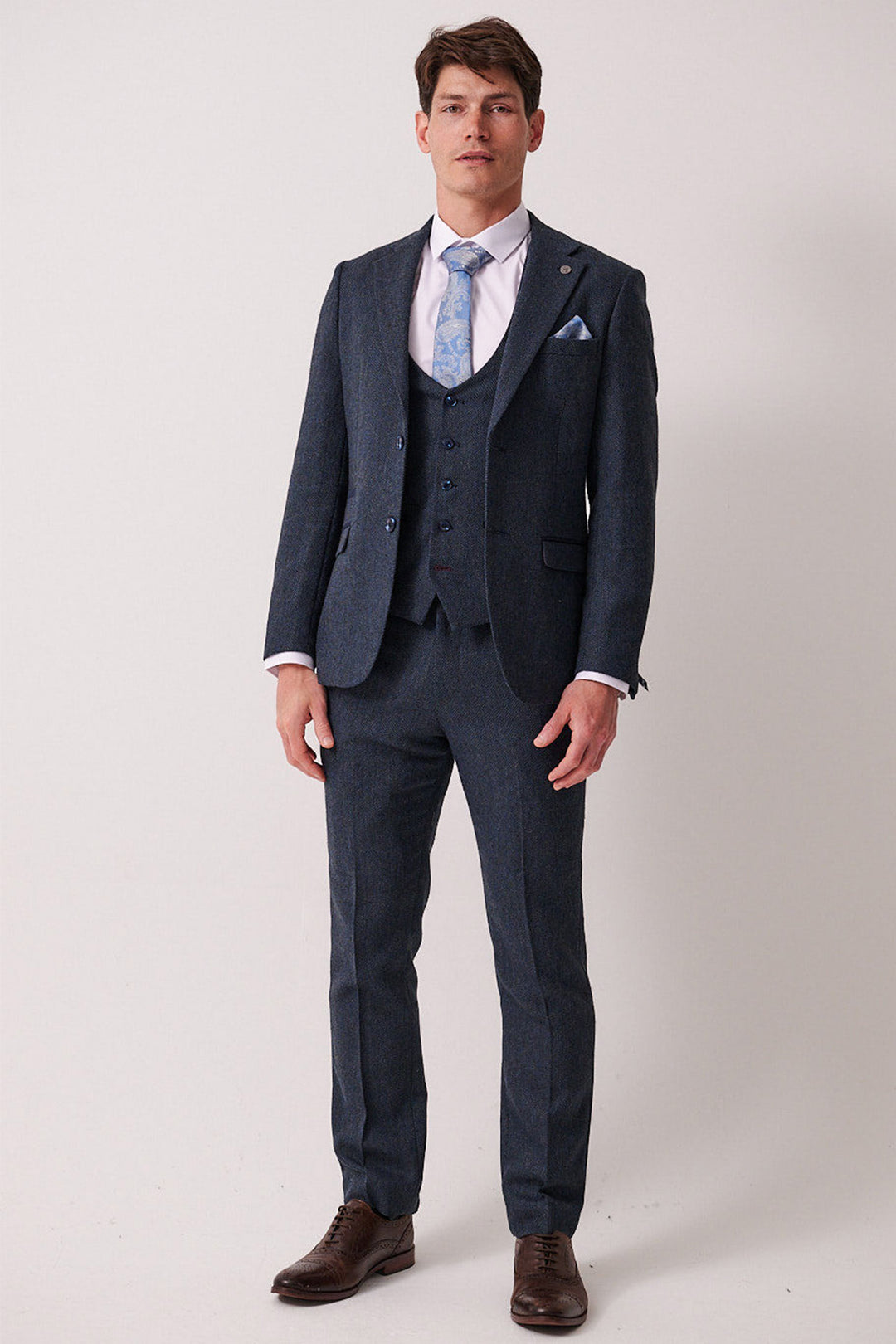 A man faces forward wearing the marcdarcy DEAN Blue Tweed Three Piece Suit with a white shirt, light blue patterned tie, and brown dress shoes against a plain light background.