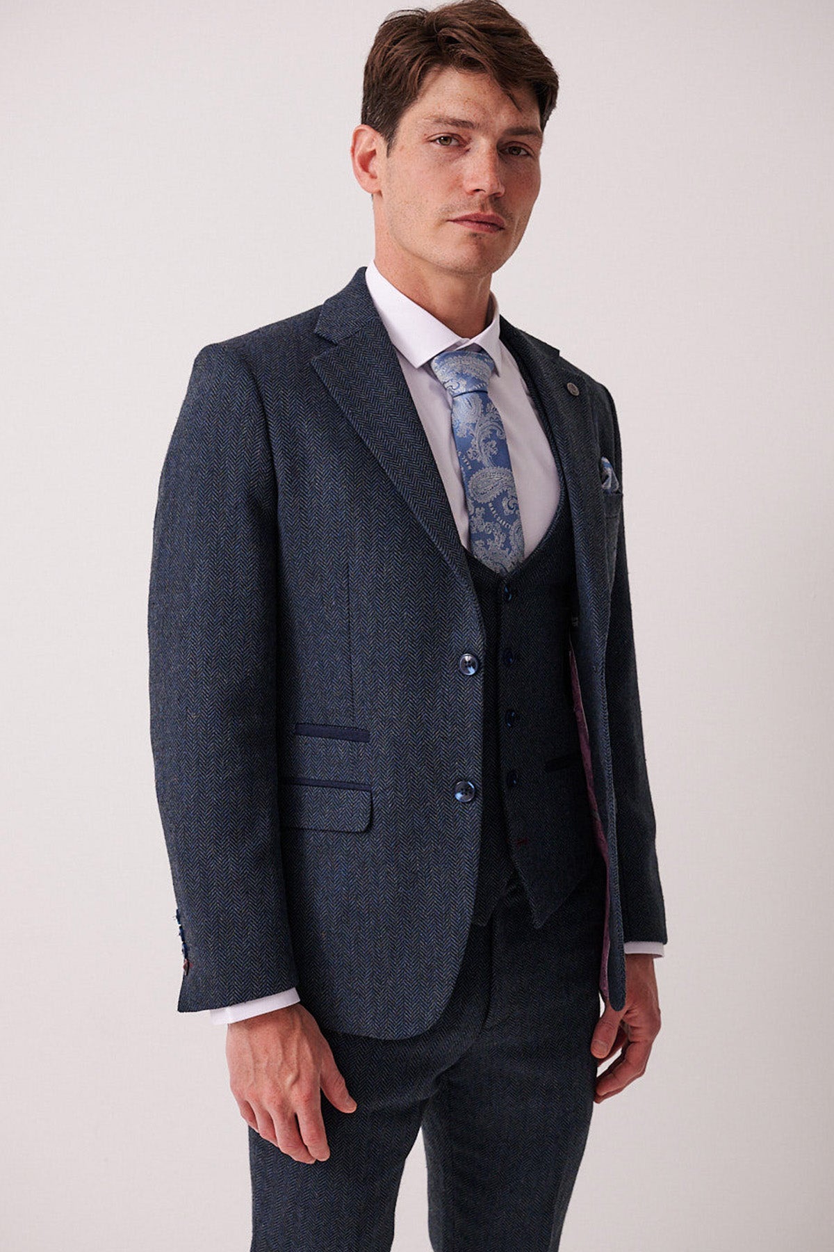 A man in the marcdarcy DEAN Blue Tweed Three Piece Suit, paired with a white dress shirt and patterned blue tie, stands confidently against a plain light background, looking at the camera.