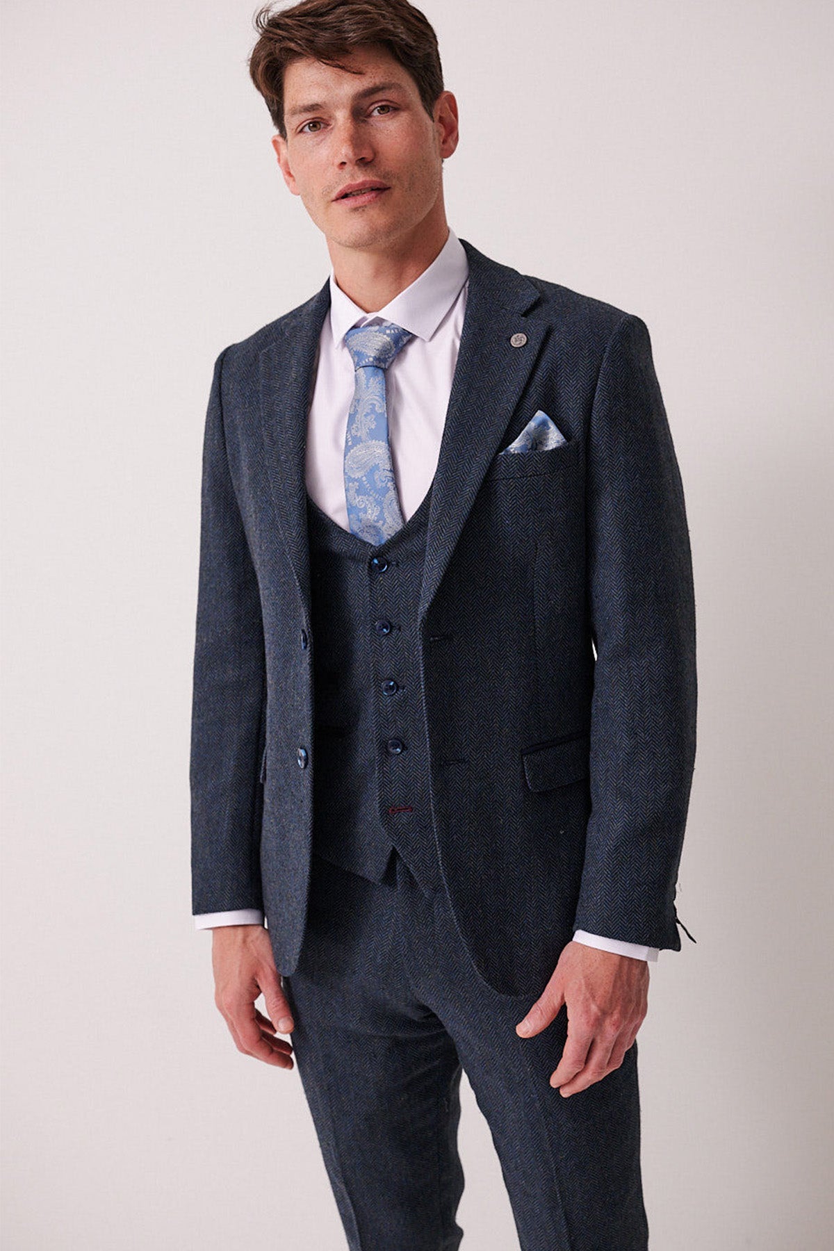 A man confidently faces the camera in a marcdarcy DEAN Blue Tweed Blazer, styled as part of a three-piece suit with a white shirt and light blue patterned tie against a plain light background.