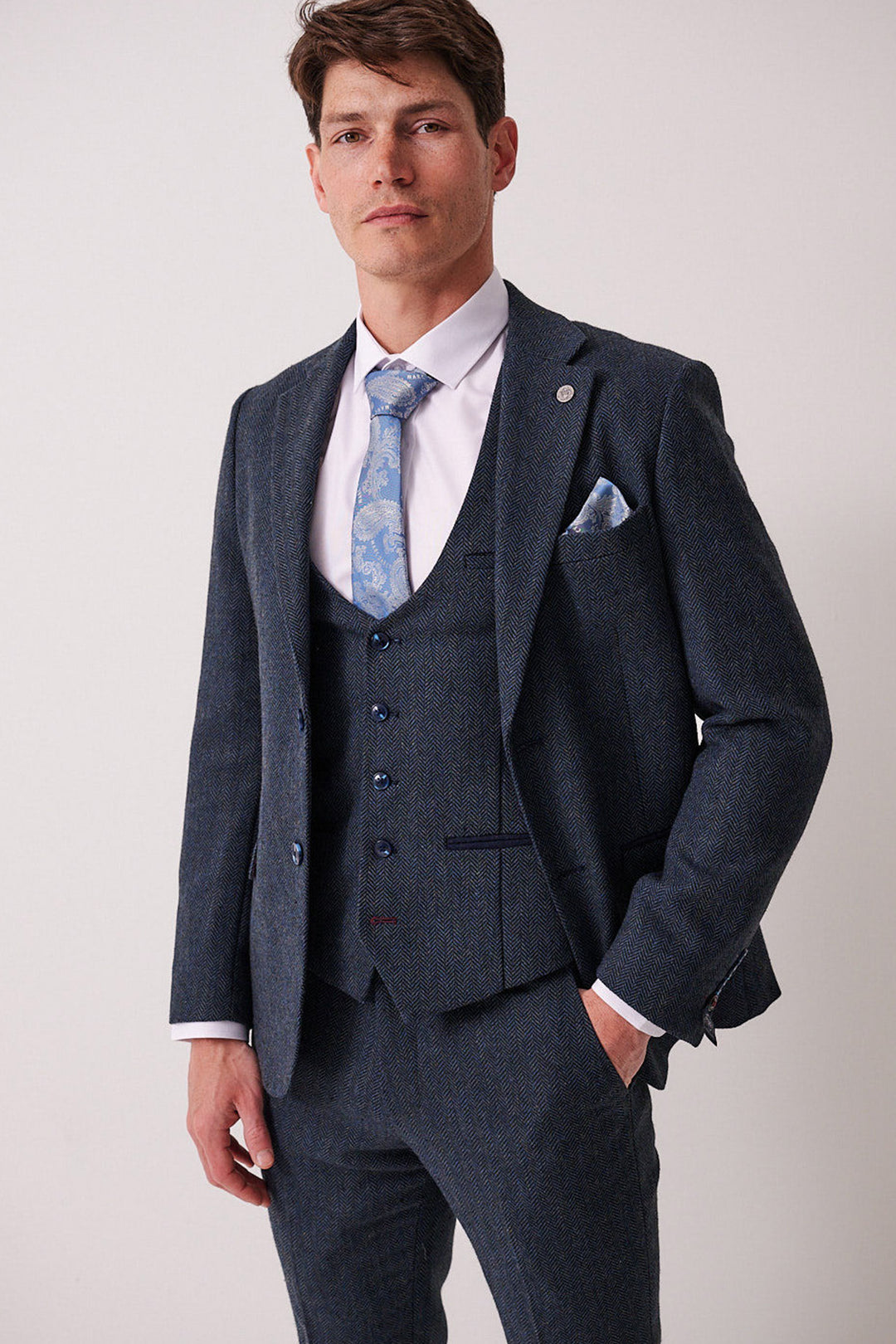 A man confidently faces the camera, wearing the marcdarcy DEAN Blue Tweed Blazer, styled with a light blue patterned tie and pocket square against a plain, light background.