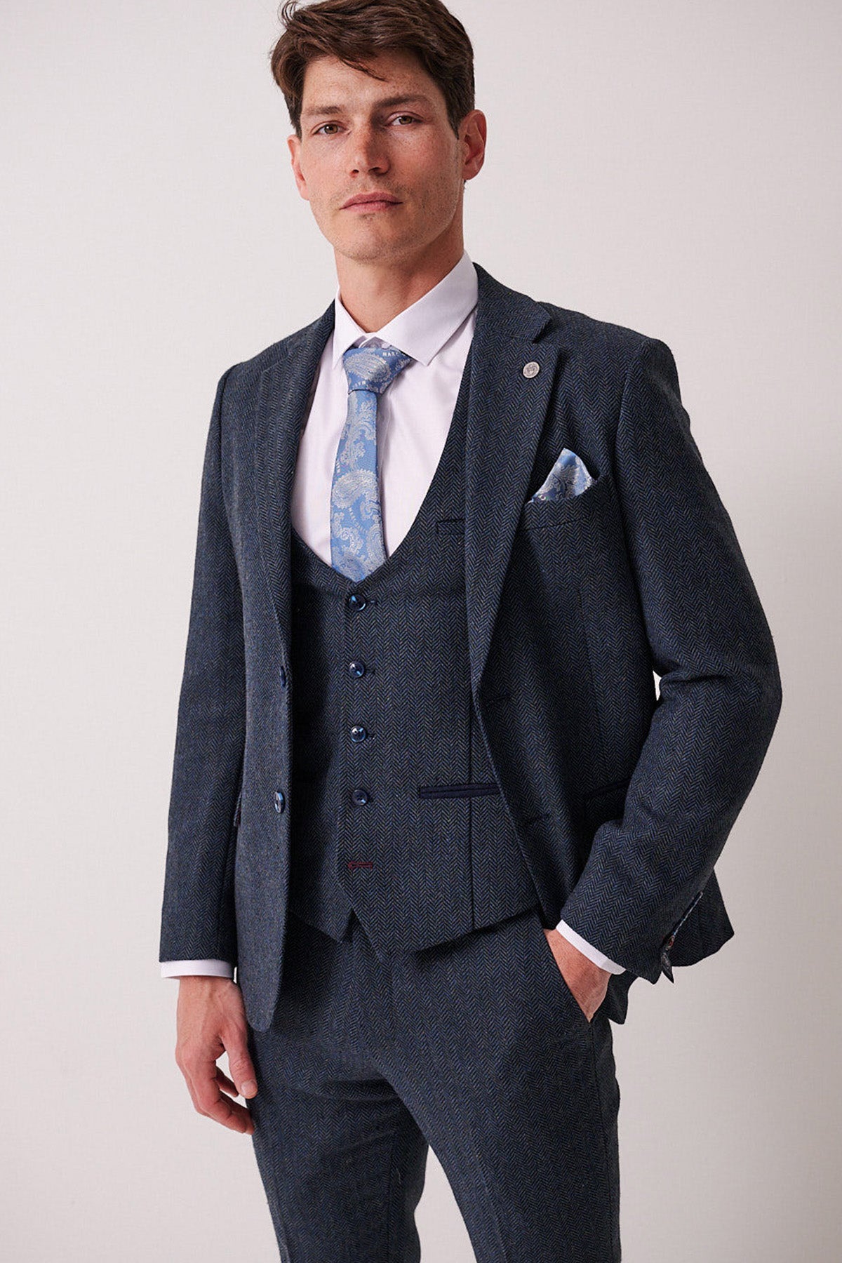 A man confidently faces the camera, wearing the marcdarcy DEAN Blue Tweed Blazer, styled with a light blue patterned tie and pocket square against a plain, light background.