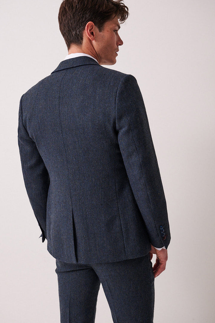 A man wears the marcdarcy DEAN Blue Tweed Three Piece Suit, standing with his back to the camera and glancing over his left shoulder. The subtly textured suit contrasts against a plain, light background.