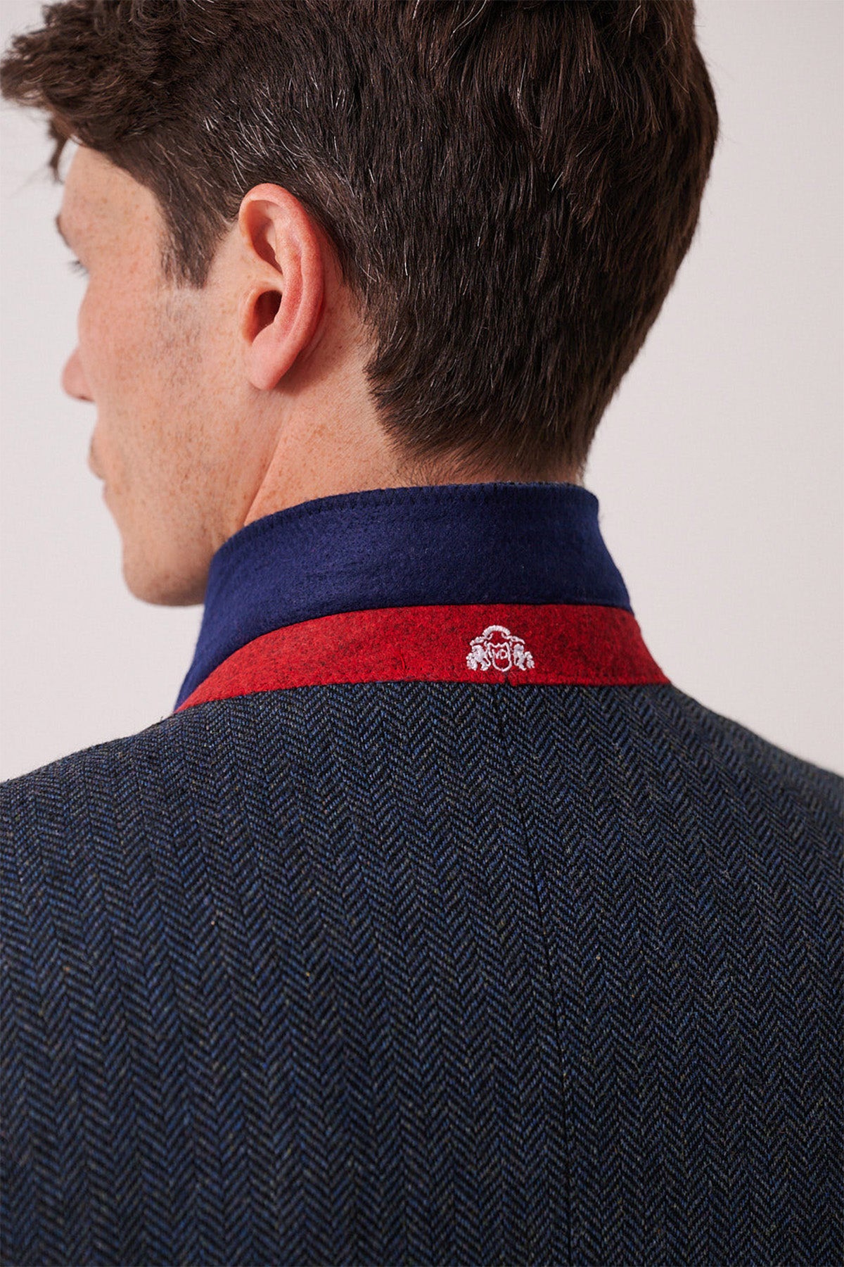 A man is seen from behind in the marcdarcy DEAN - Blue Tweed Blazer, featuring a blue and red collar with a small white embroidered design. The classic herringbone adds timeless style as he turns his head slightly left.