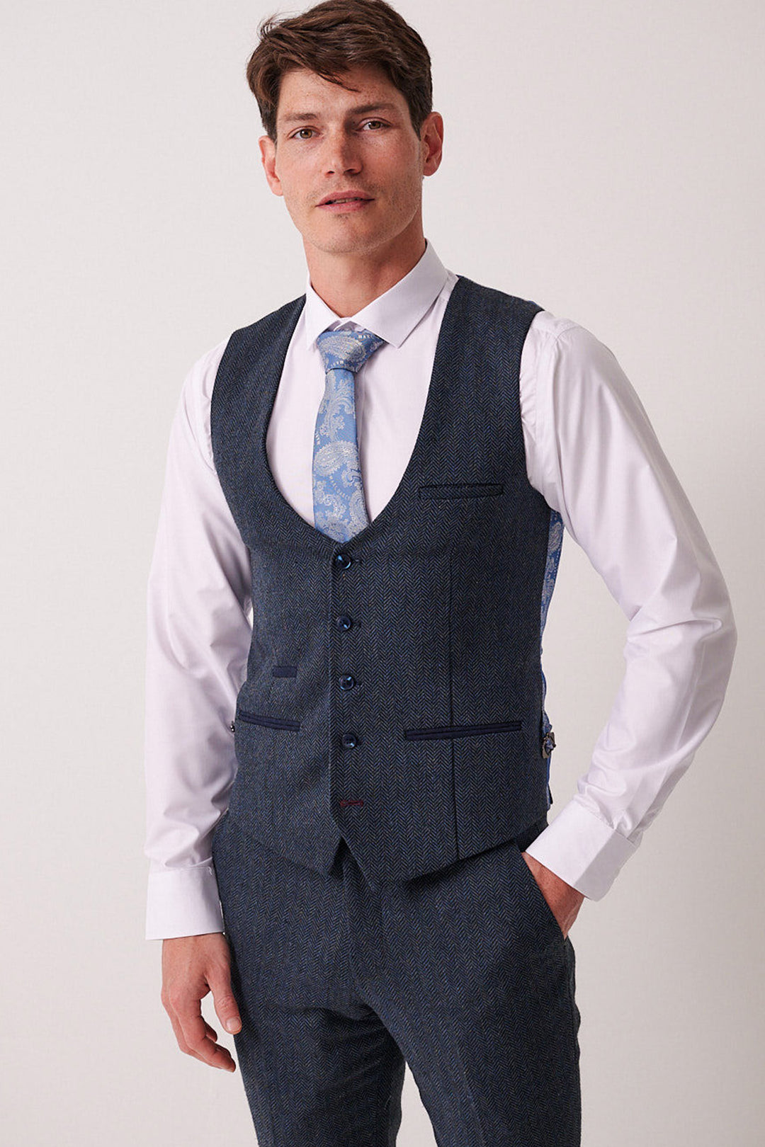 A man wears a Marc Darcy DEAN Blue Tweed Waistcoat and matching trousers, paired with a white dress shirt and light blue patterned tie, standing with one hand in his pocket against a plain light background.