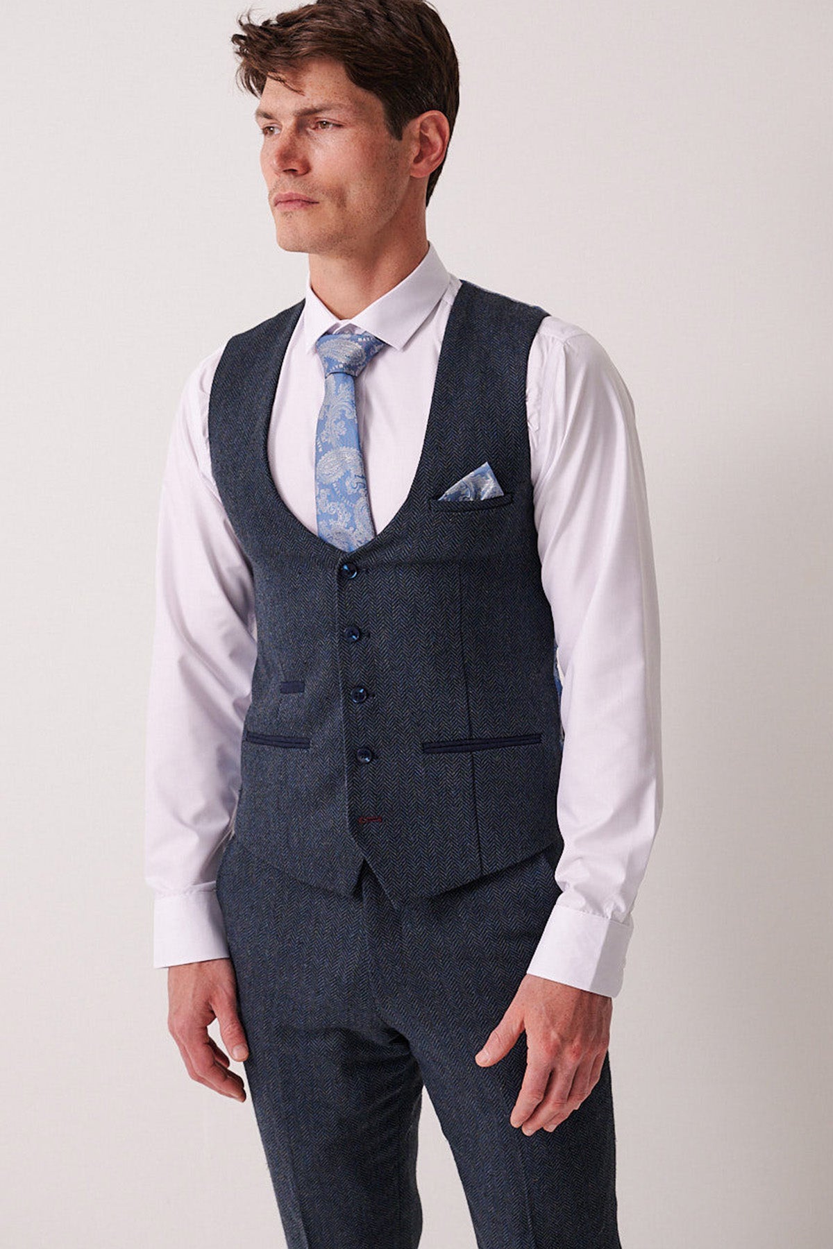 A man stands against a plain light background, looking to the side, dressed in the Marc Darcy DEAN Blue Tweed Waistcoat with matching trousers, a white dress shirt, and a blue patterned tie.